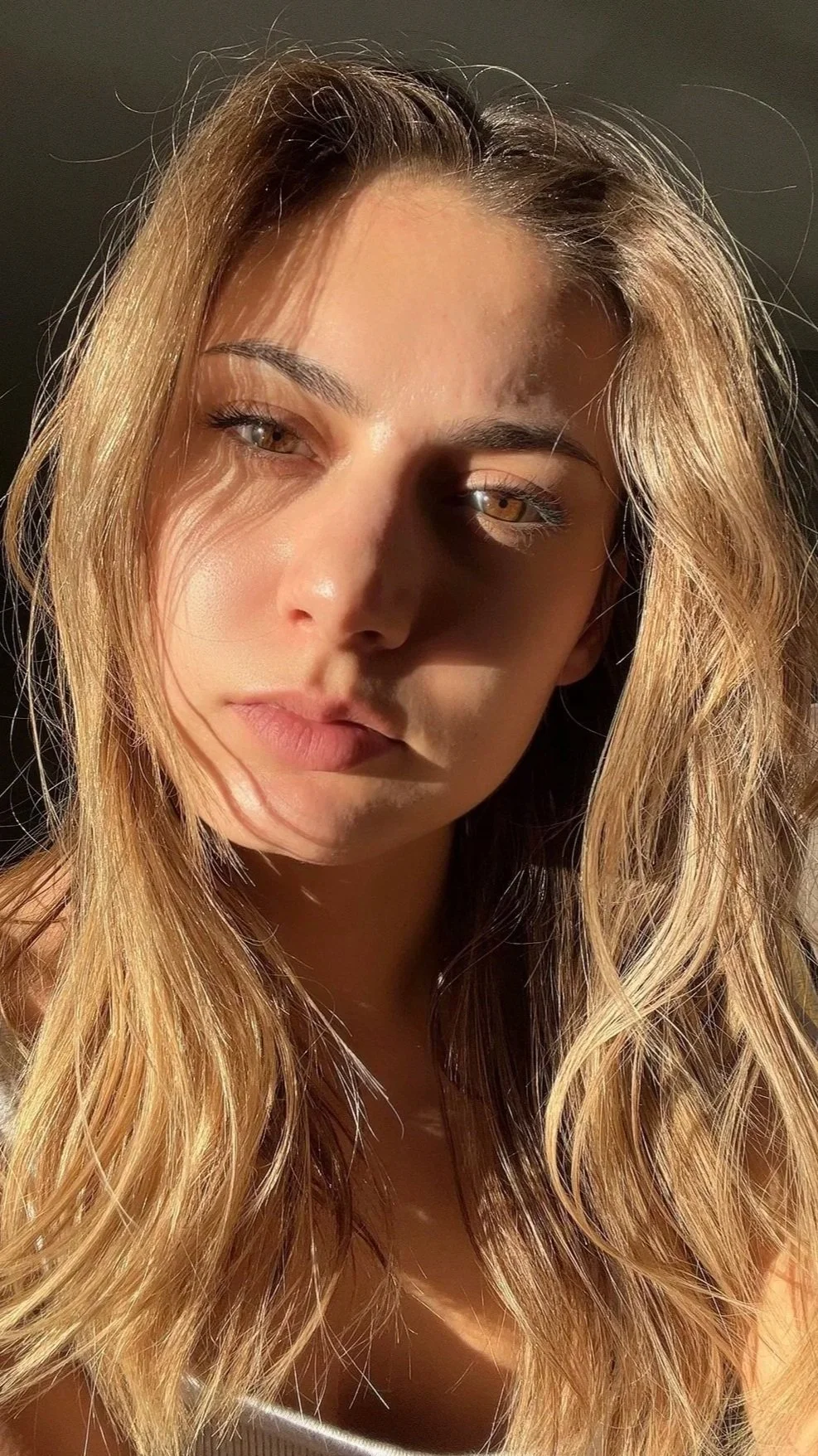 Close-up of a young woman with long, wavy blonde hair, partially shadowed face, neutral expression, and subtle makeup, lit by warm sunlight.