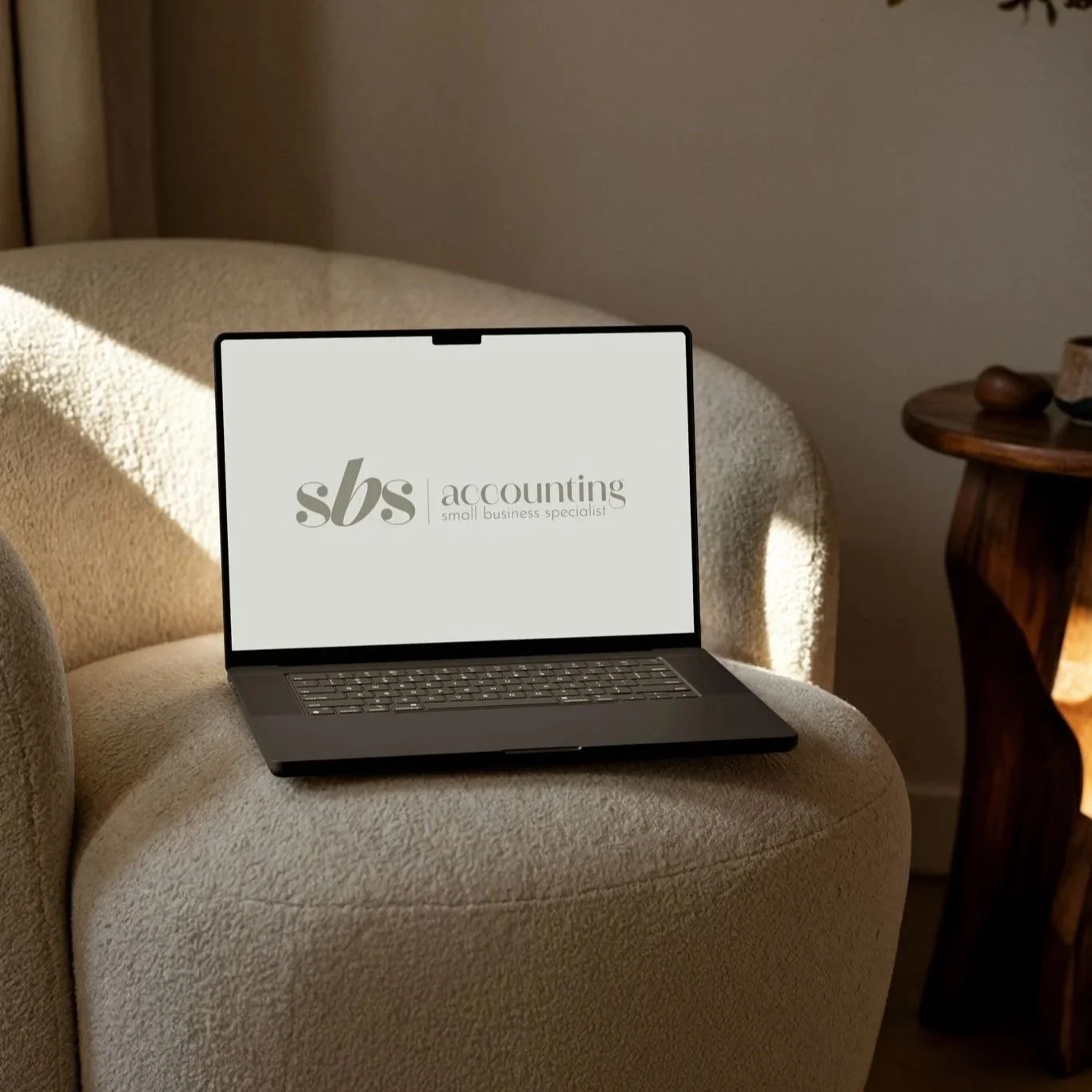 Laptop on beige upholstered chair displaying the logo of sb s accounting, small business specialist, with sunlight streaming in.