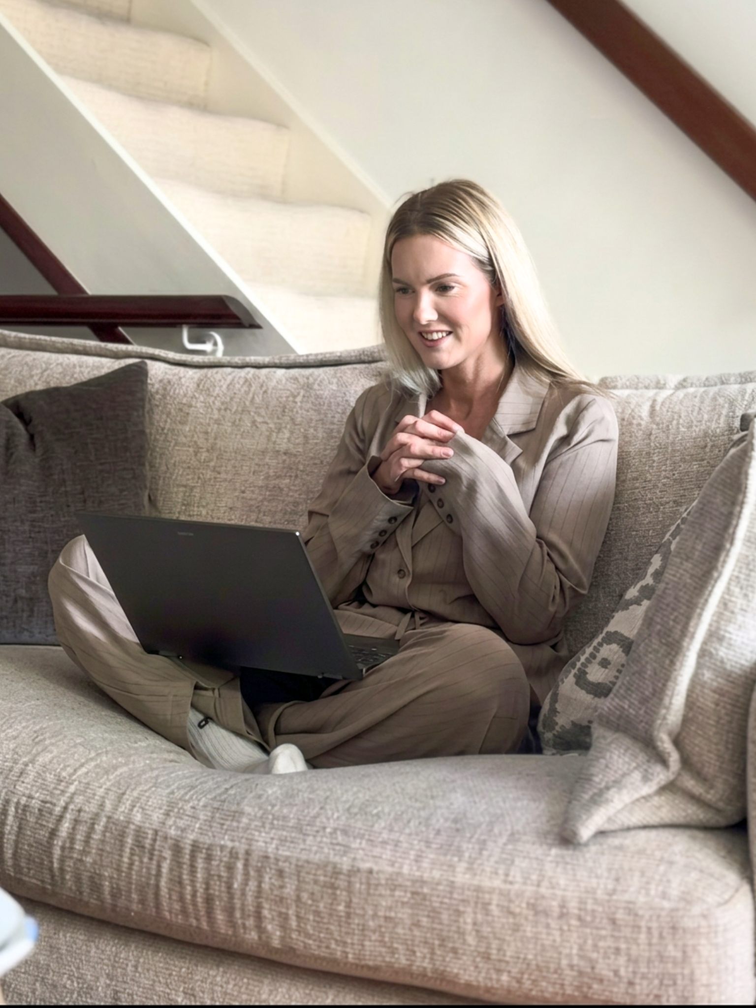 A young woman with blonde hair sitting cross-legged on a beige sofa, looking at a laptop, in a home with a staircase in the background.