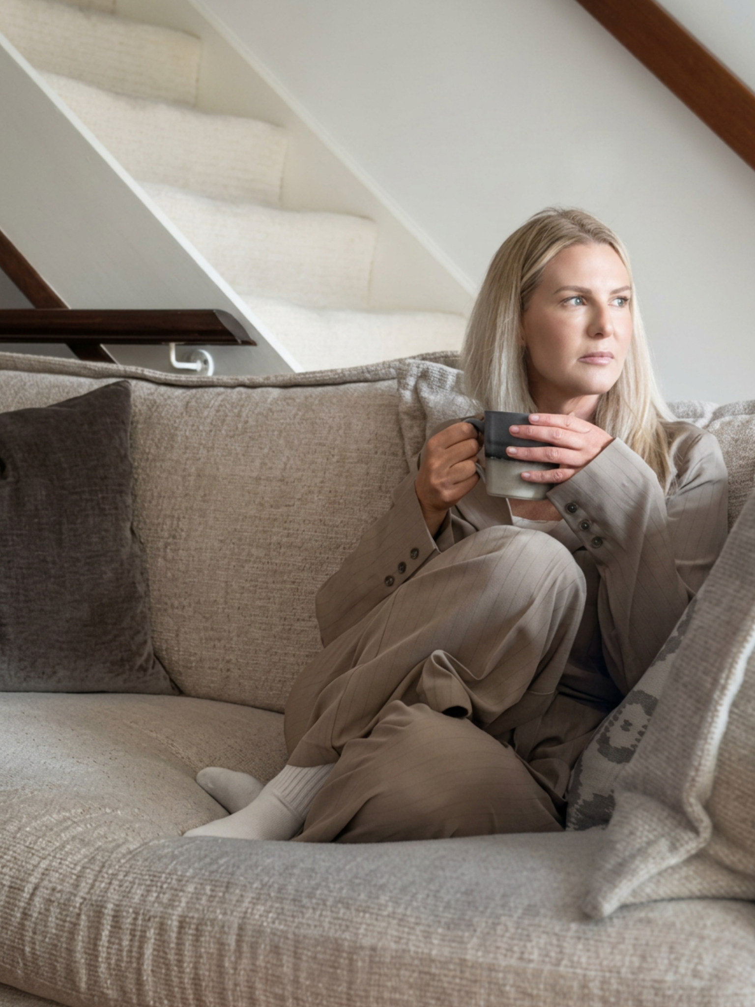 A woman with long blonde hair seated on a beige sofa, holding a mug, with a neutral facial expression, in a room with a sloped ceiling and light-colored walls.