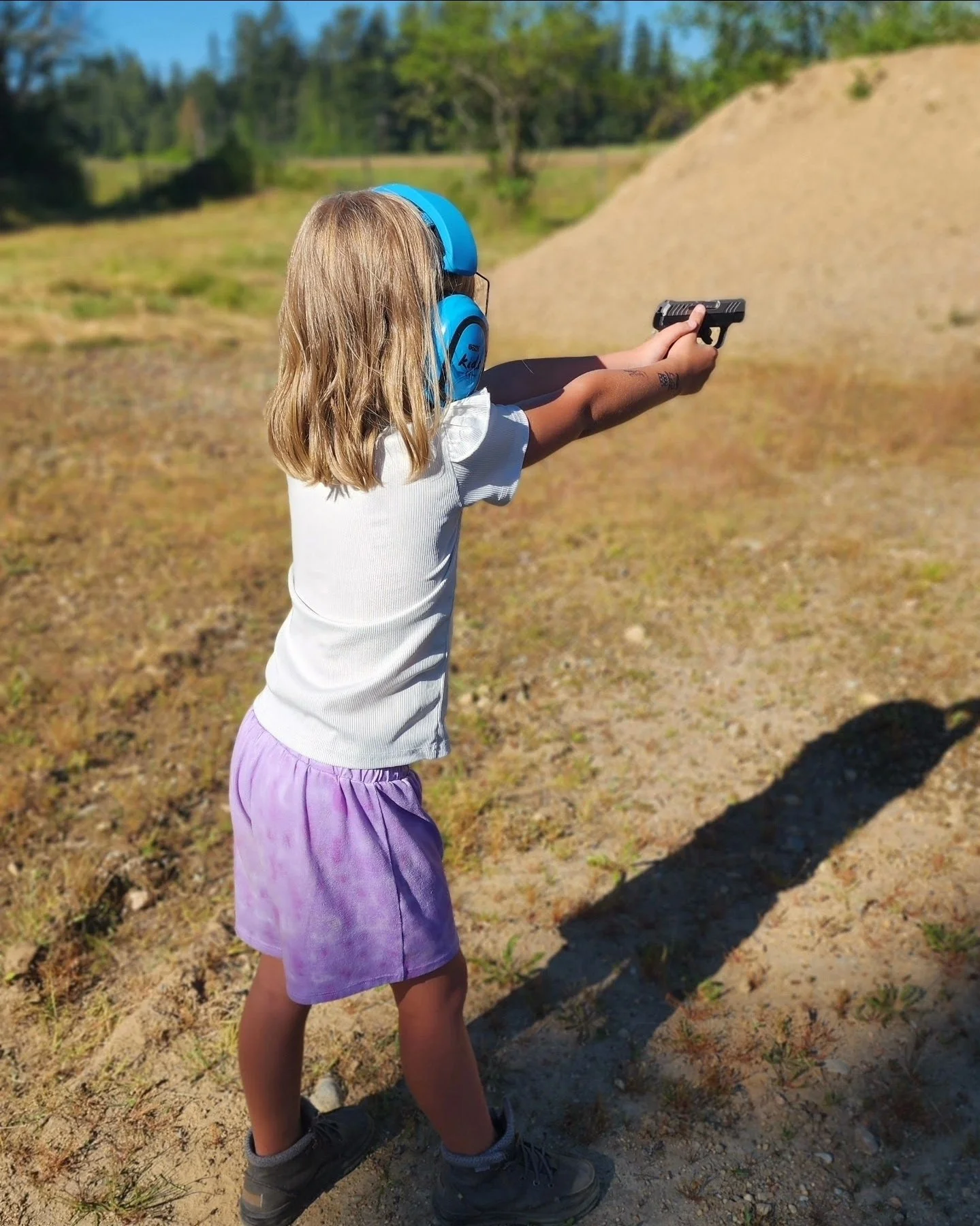 A young girl with blonde hair, wearing a white t-shirt, purple skirt, and grey boots, is standing outdoors at shooting range, aiming a small handgun, with blue noise-canceling headphones on.