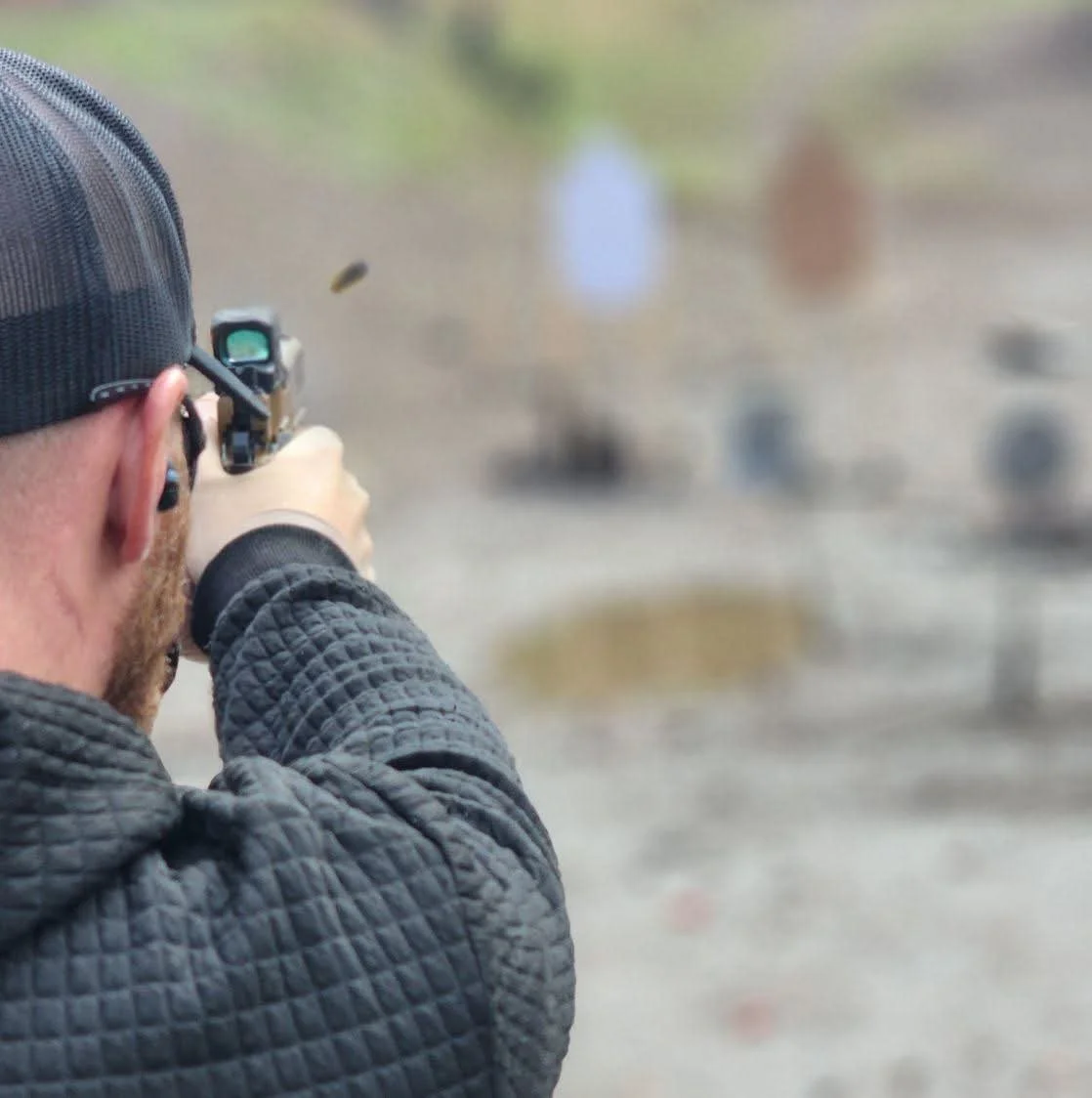 A person aiming a handgun with a scope, wearing a black cap and dark quilted jacket, at an outdoor shooting range with blurred targets in the background.