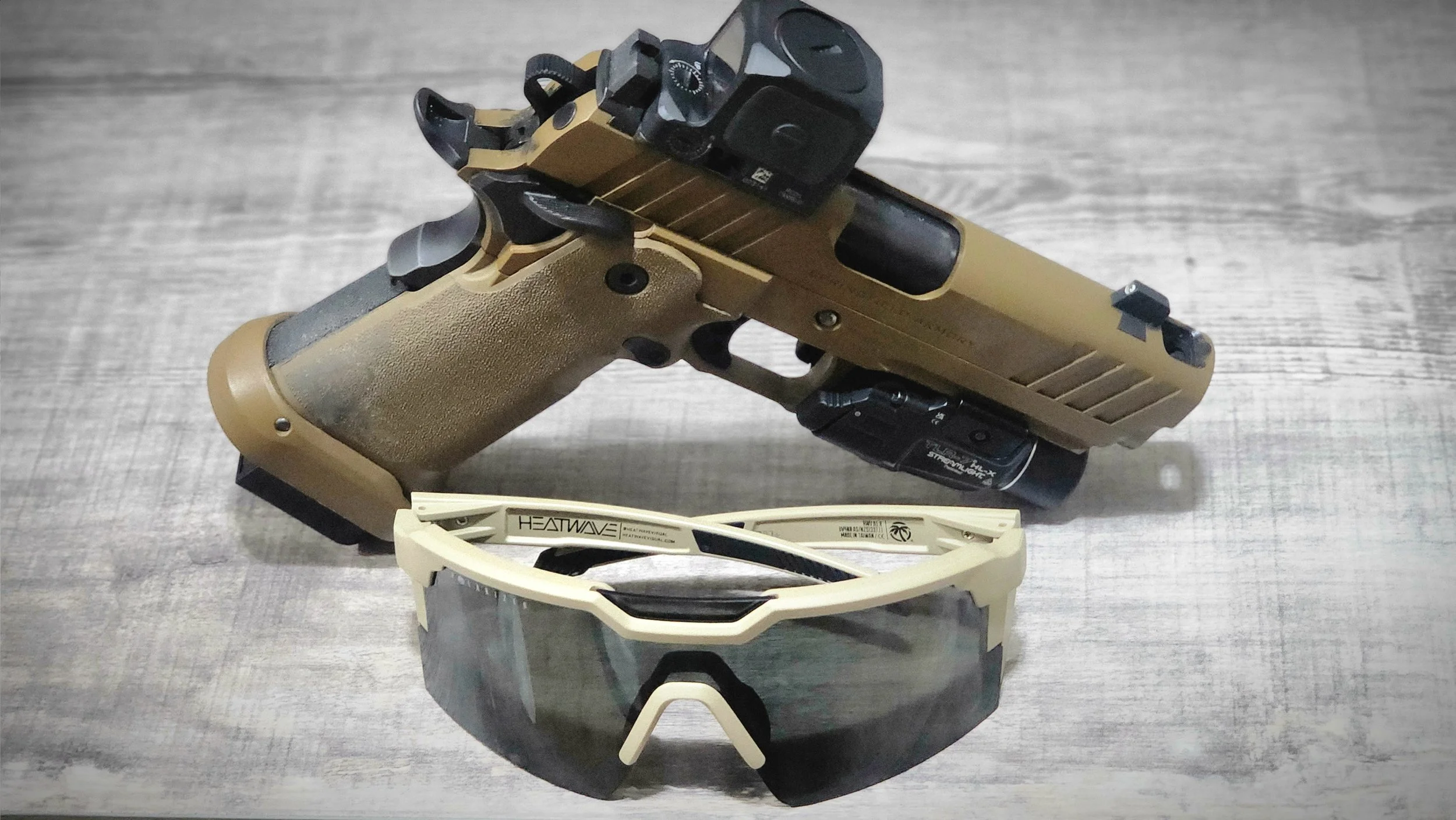 A tan and black handgun with a mounted scope, placed on a gray wooden surface, with a pair of white sports sunglasses with black lenses and a logo on the arm in front of it.