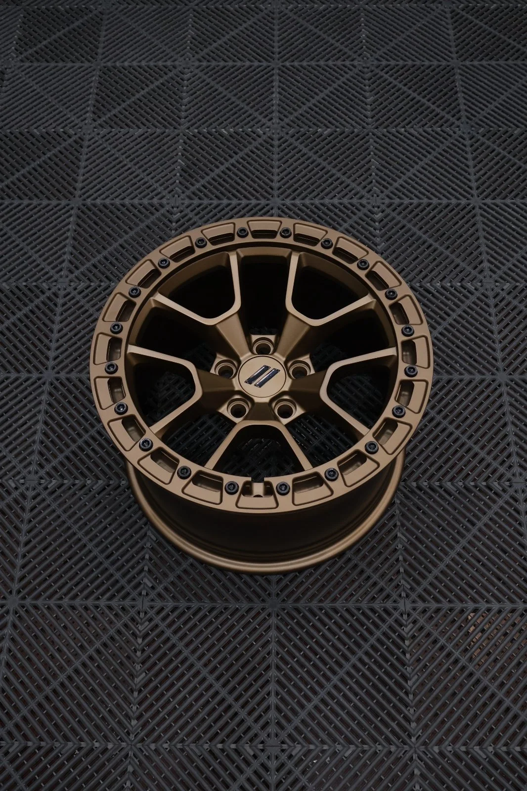 A bronze-colored alloy wheel rim with a modern, angular design is placed on a black textured rubber mat with a diamond pattern.