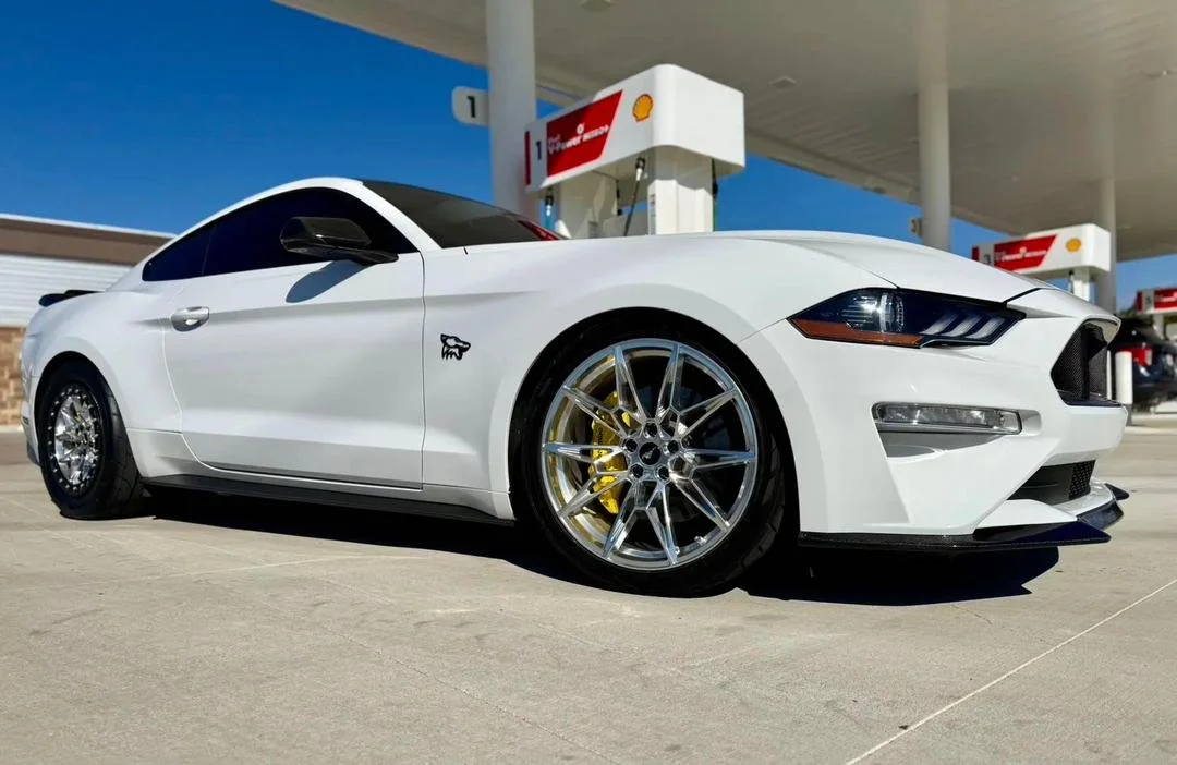 White sports car parked at a Shell gas station with a low-angle view, showing custom wheels and a black emblem of a falcon on the door.