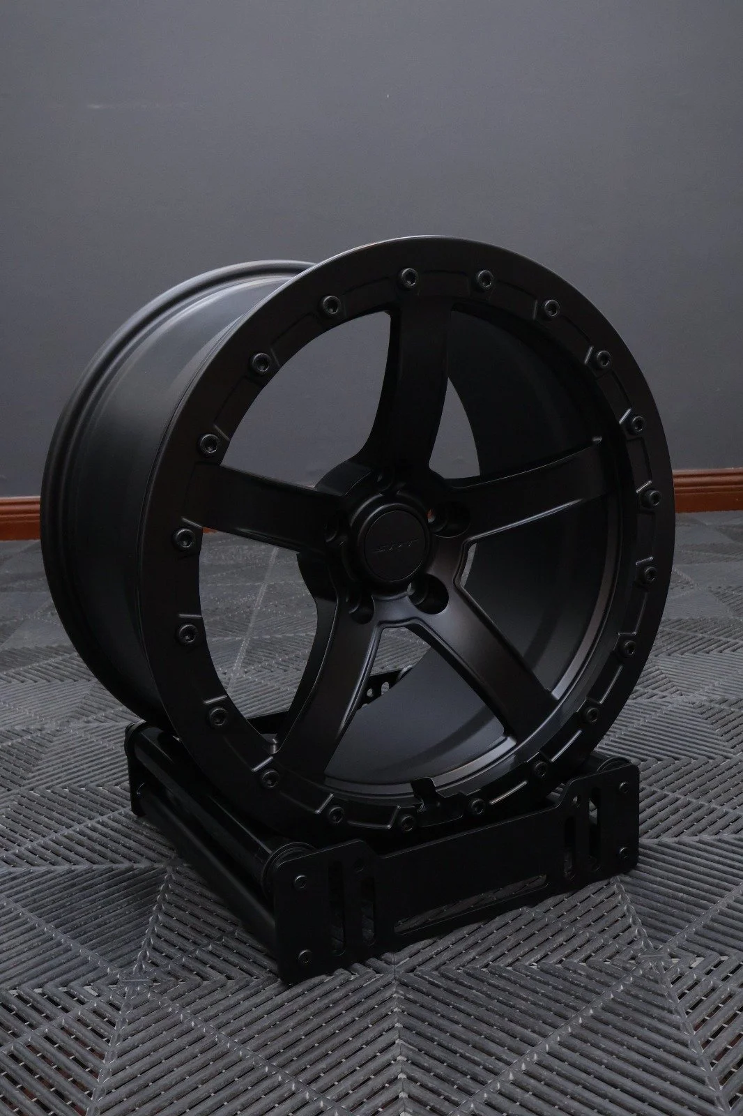 A black, custom-designed wheel rim mounted on a display stand, placed on a textured floor with a plain gray wall background.