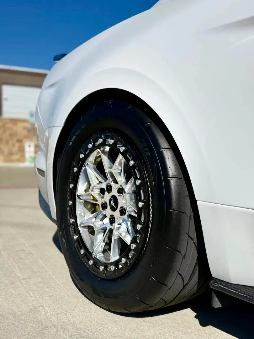 Close-up of a white sports car wheel with a black tire and silver rim, parked on a concrete surface under a clear blue sky.