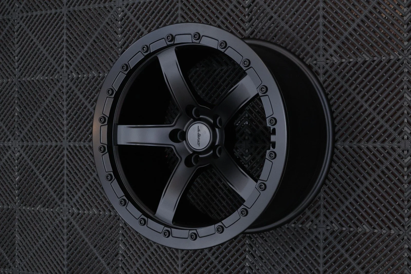 A black aftermarket car wheel rim with a multi-spoke design, resting on a textured black rubber mat.