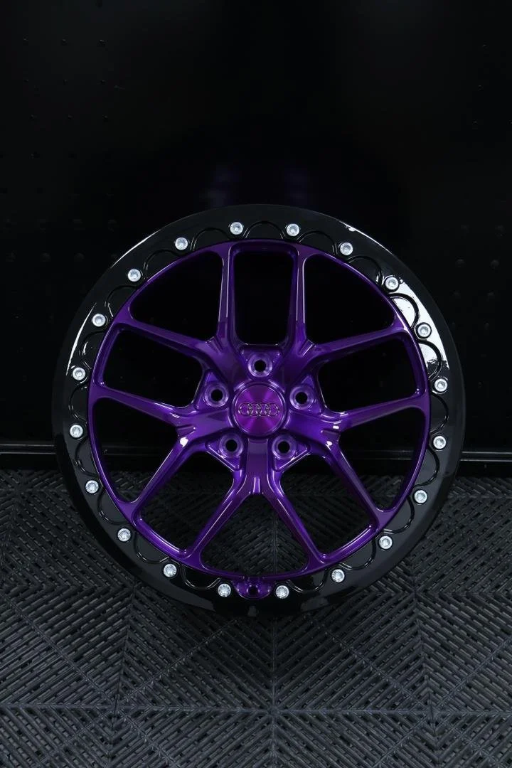 A purple Audi wheel rim with a black outer edge and silver bolts, placed on a black textured surface.