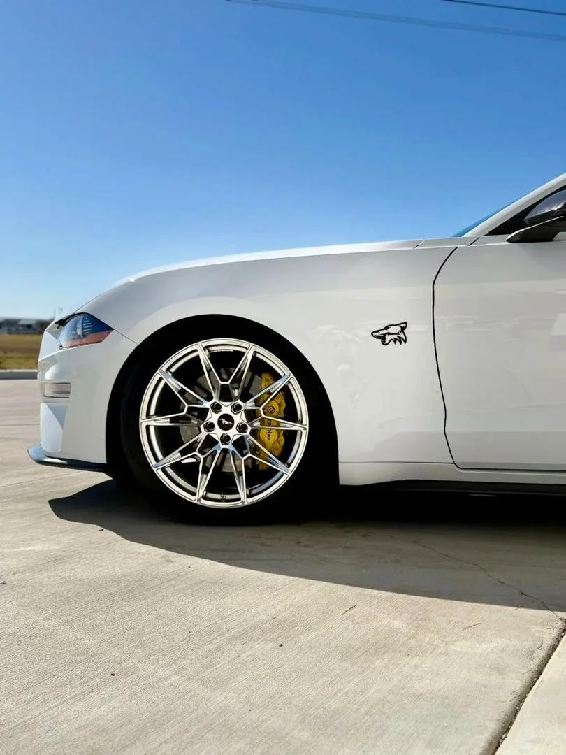 Close-up of a white sports car parked on a concrete surface, showing the front wheel with a yellow brake caliper and part of the car's body with a small logo of an animal head.