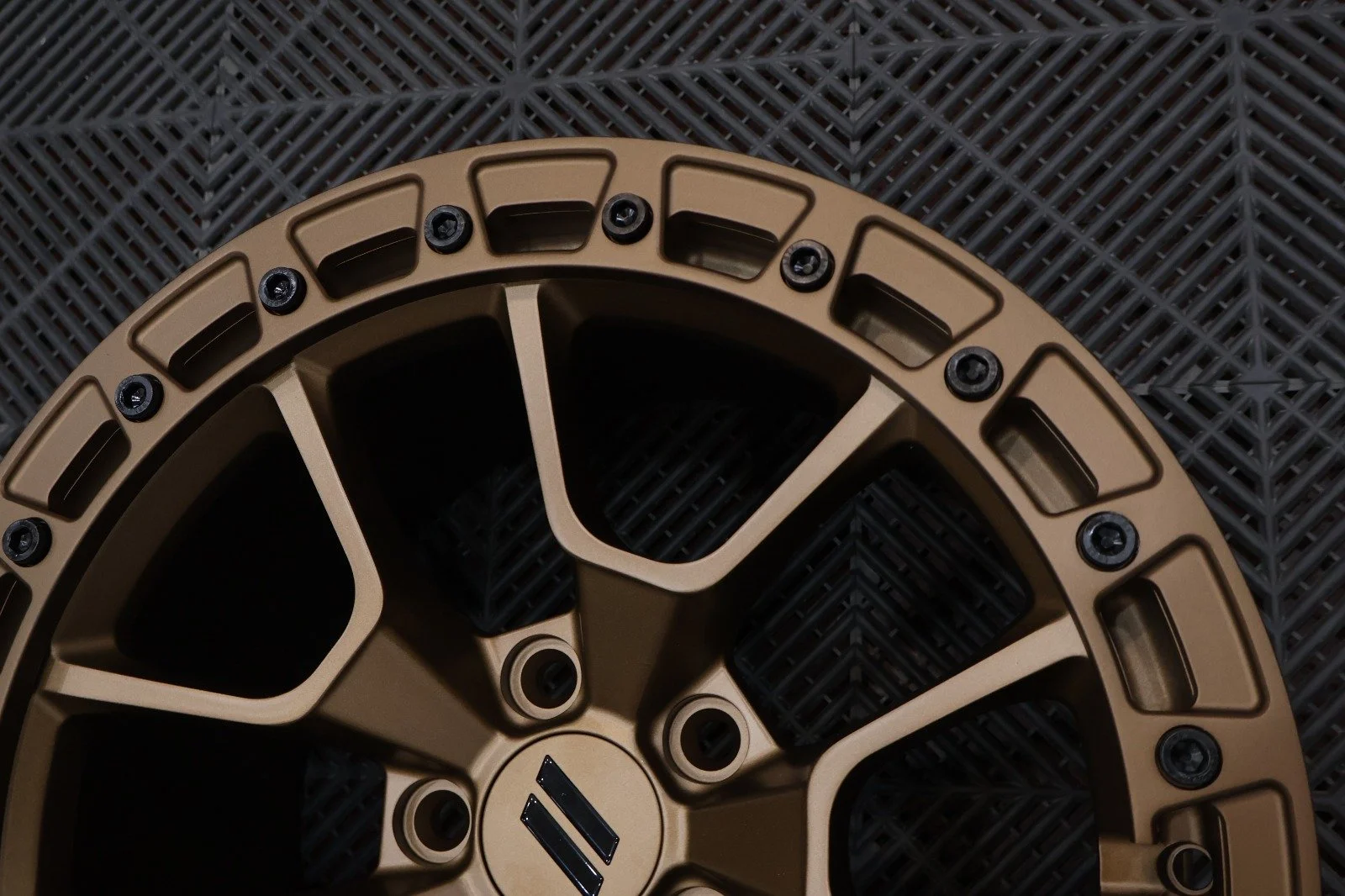 Close-up of a bronze-colored, multi-spoke, round metal wheel with black screws, resting on a black plastic grid surface.