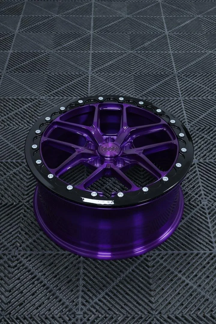 A purple aftermarket wheel rim with a black lip, placed on a black textured surface.