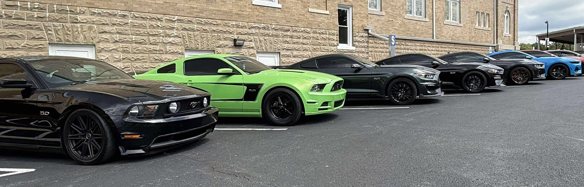 Line of six parked Ford Mustangs in various colors including black, green, gray, and blue in a parking lot next to a brick building.