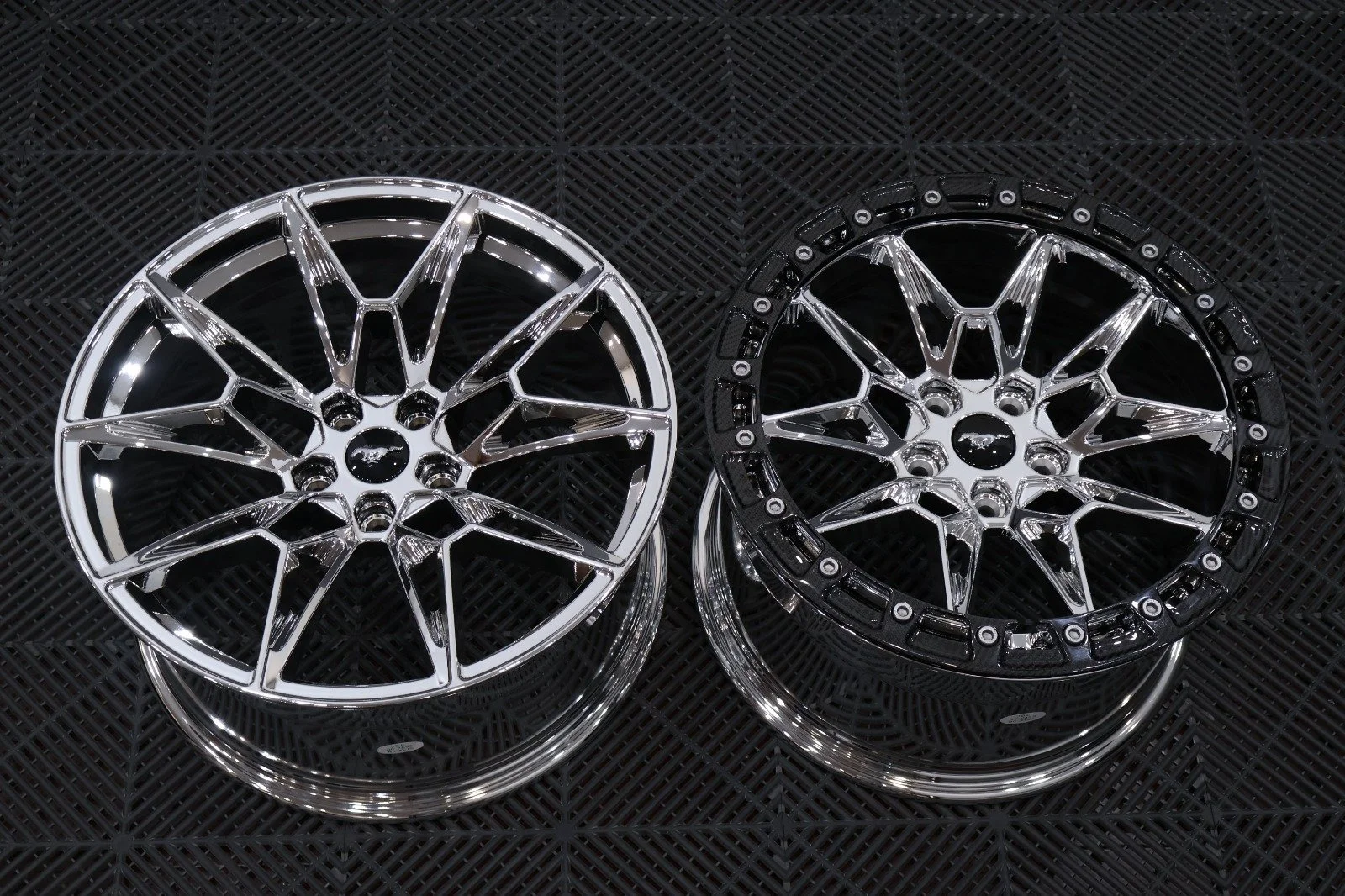 Two racing wheels with intricate spoke designs, one silver and one black, displayed on a black textured surface.