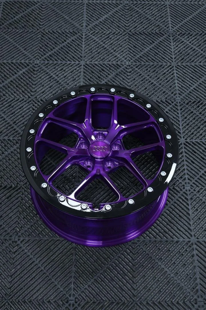 A purple and black Audi wheel rim with a multi-spoke design, placed on a textured black surface.