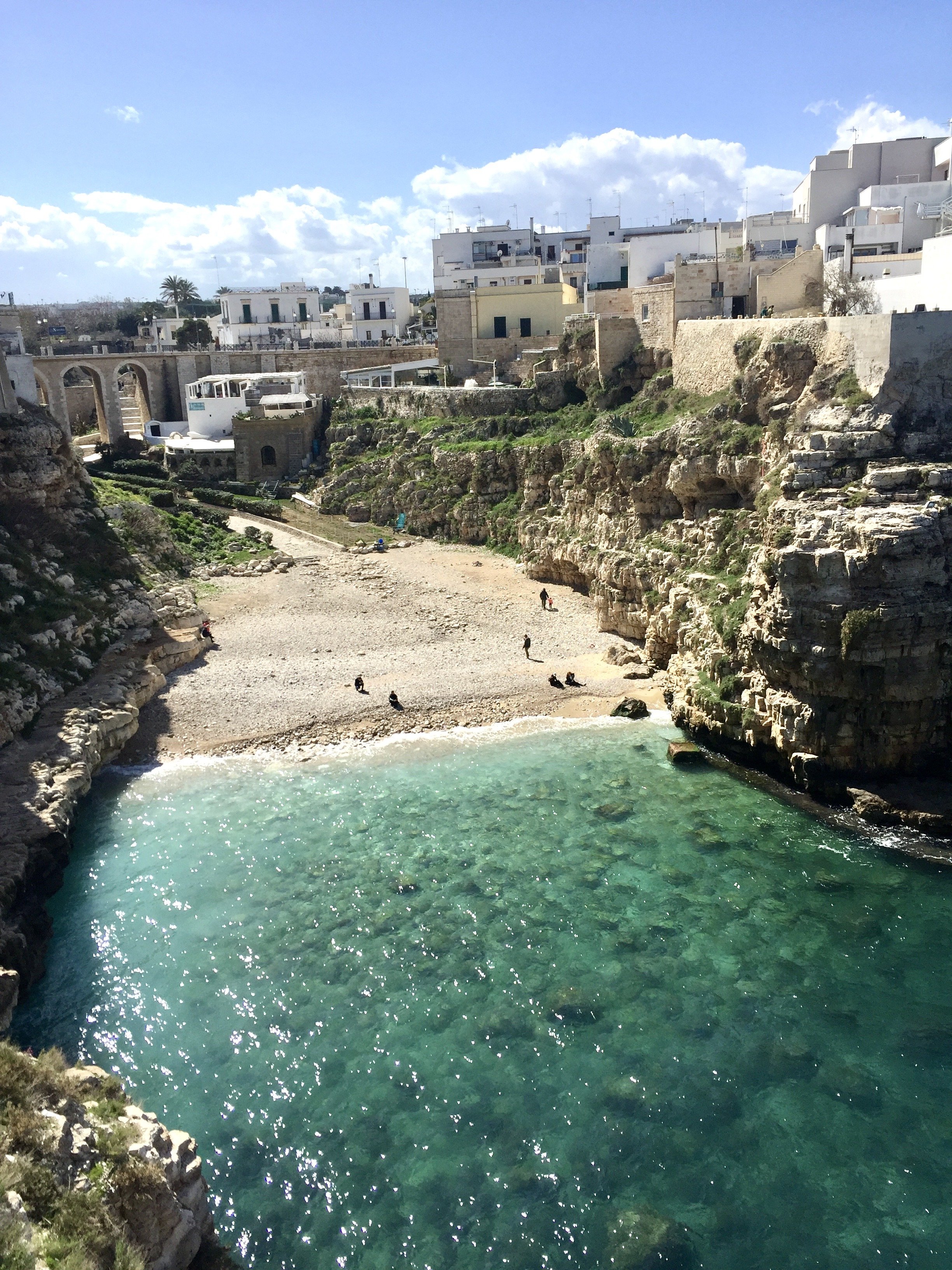 A small beach in Polignano a Mare with turquoise water bordered by rocky cliffs, nearby white buildings, and a partly cloudy sky.
