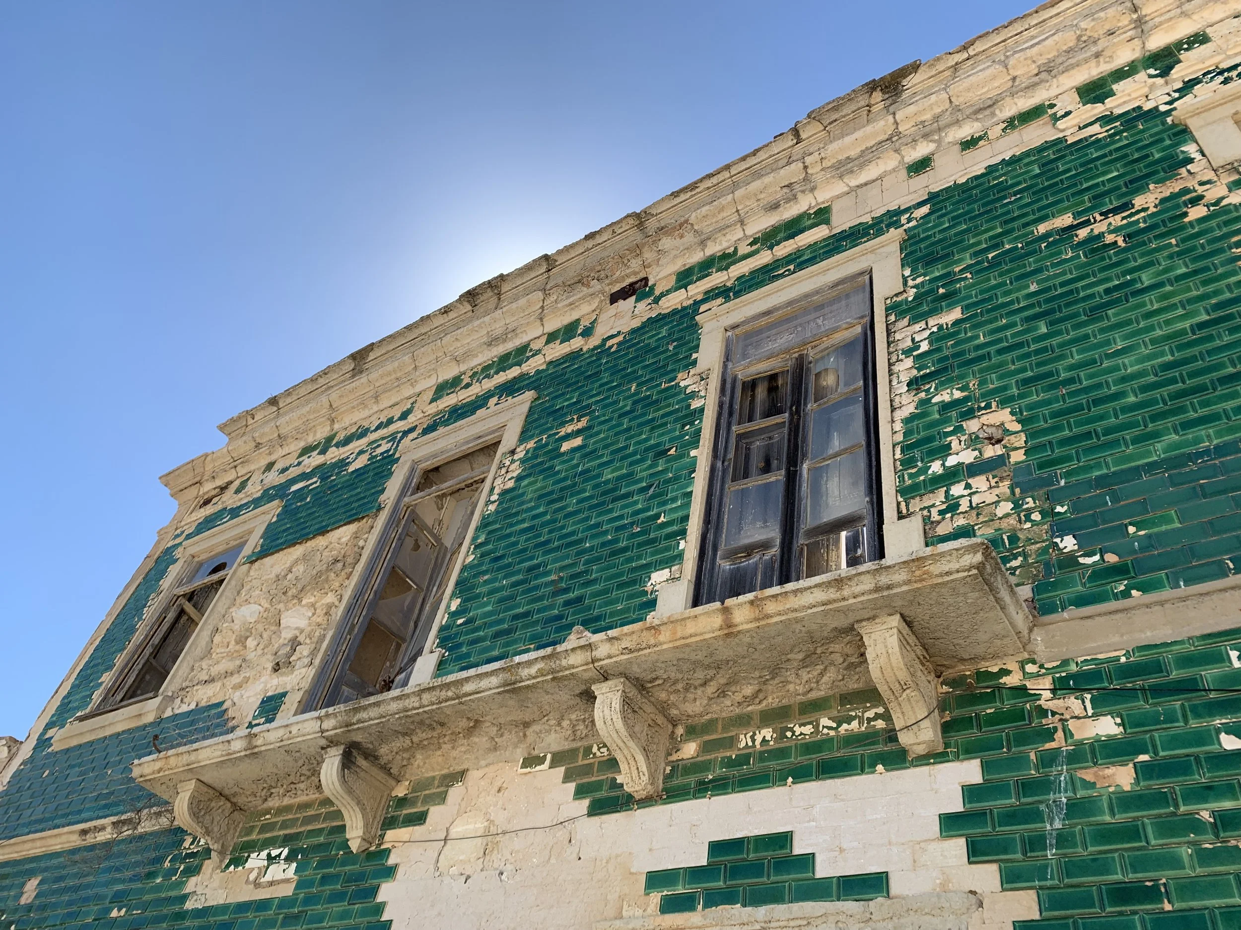 Old building with green tiles partially peeling, two tall windows with weathered shutters, stone balcony, under a clear blue sky.