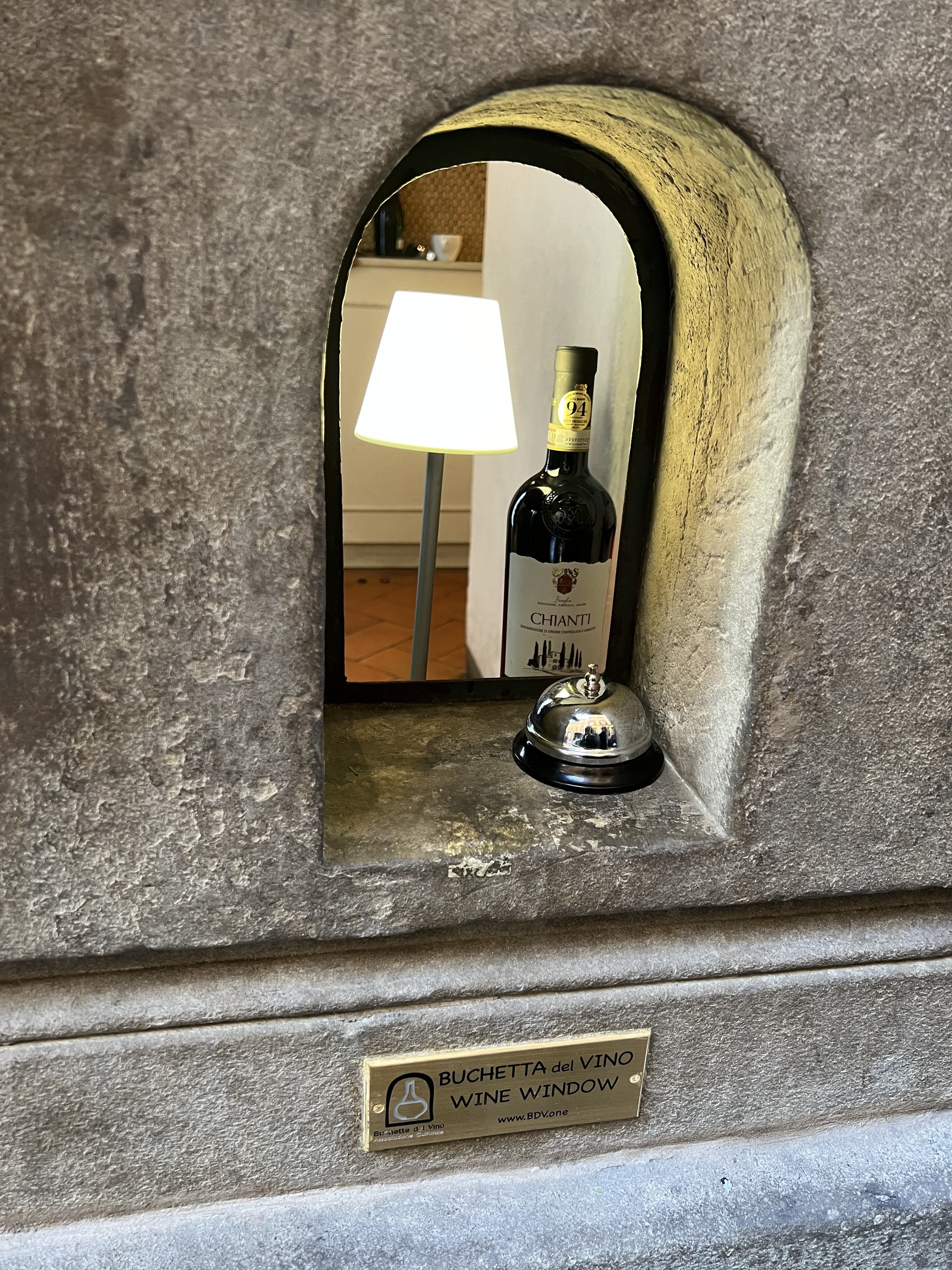 A small wine window in Florence with a metal bell, a bottle of Chianti wine, and a lamp.