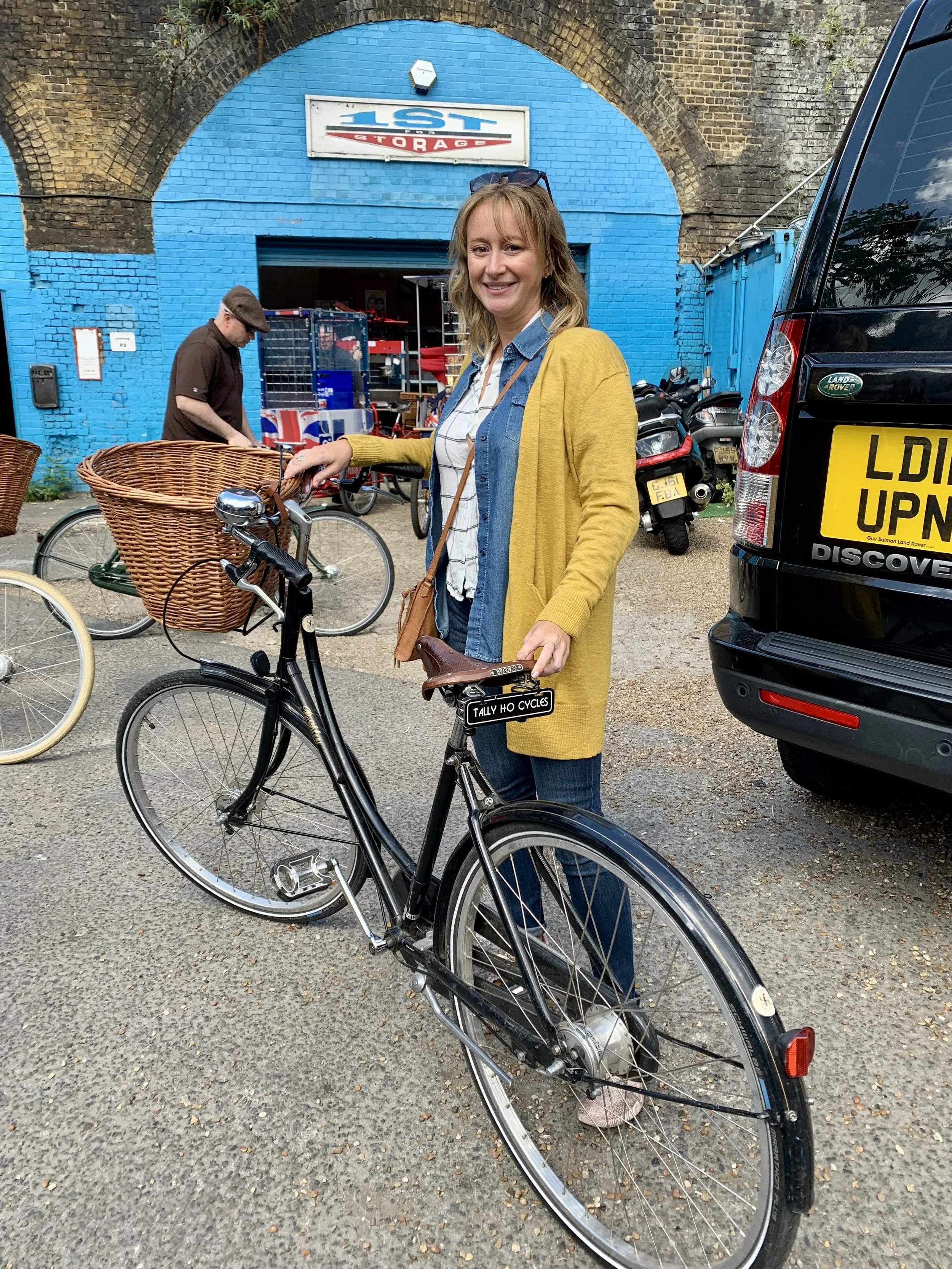 A woman in a yellow cardigan, blue jeans, and a plaid shirt stands next to a black bicycle with a wicker basket attached to the front, in front of a bright blue building.