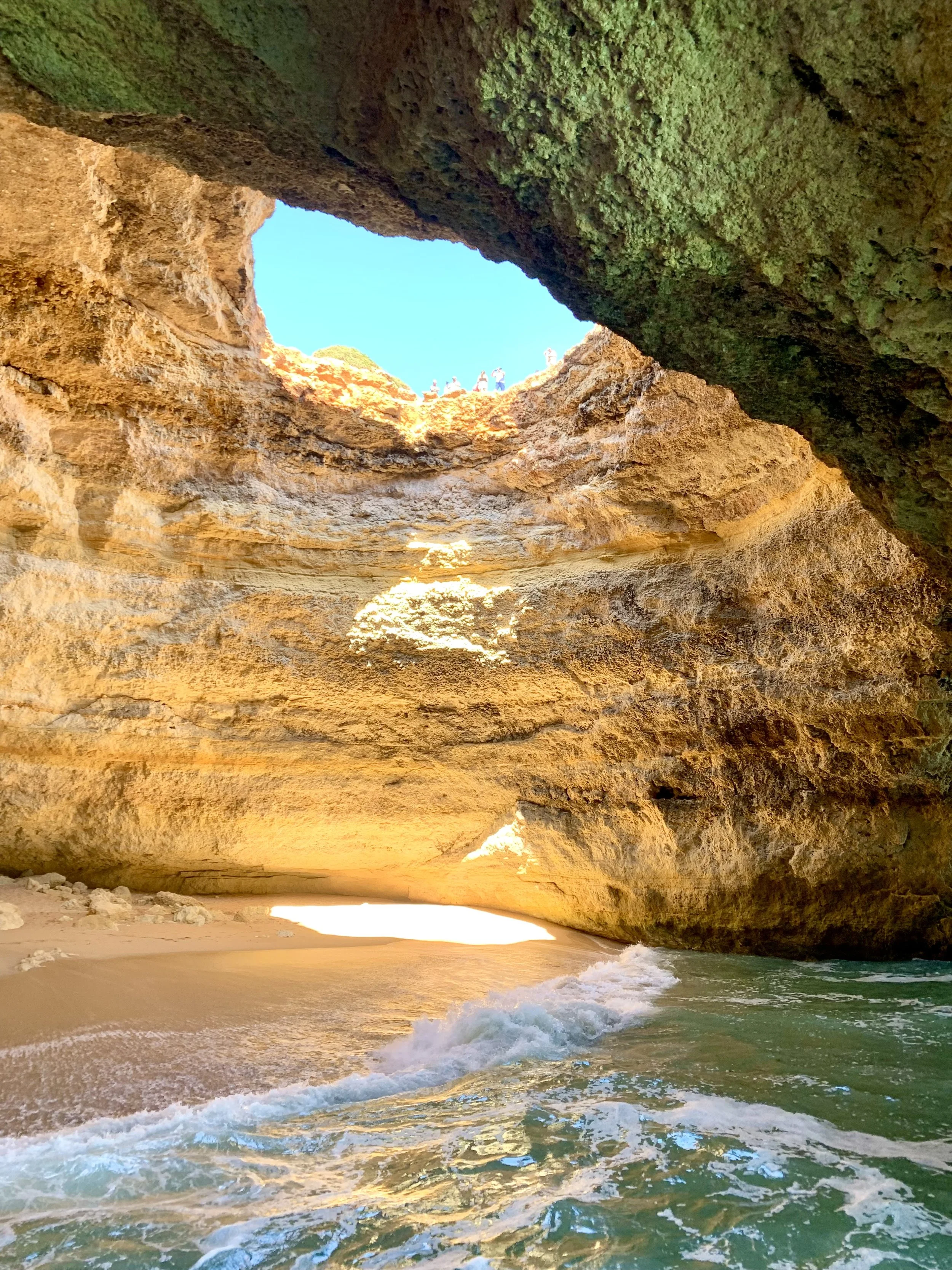 View of the Benagil Sea Cave with a sandy beach, ocean waves, and rocky cliffs with a natural hole at the top through which the blue sky is visible.