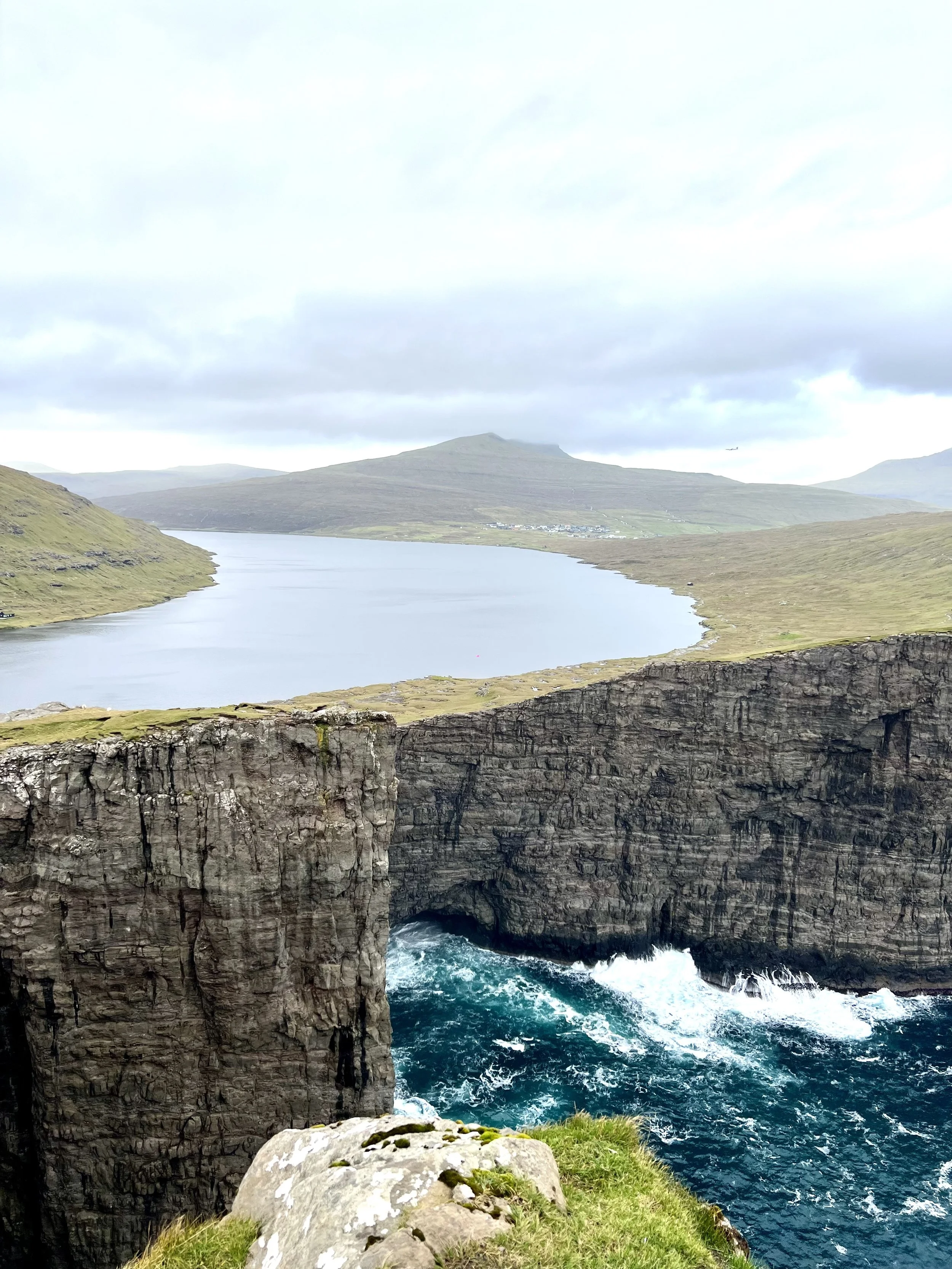 View of The Floating Lake in the Faroe Islands, a rocky sea cliff with a natural arch, overlooking a body of water with nearby green rolling hills and mountains under a cloudy sky.