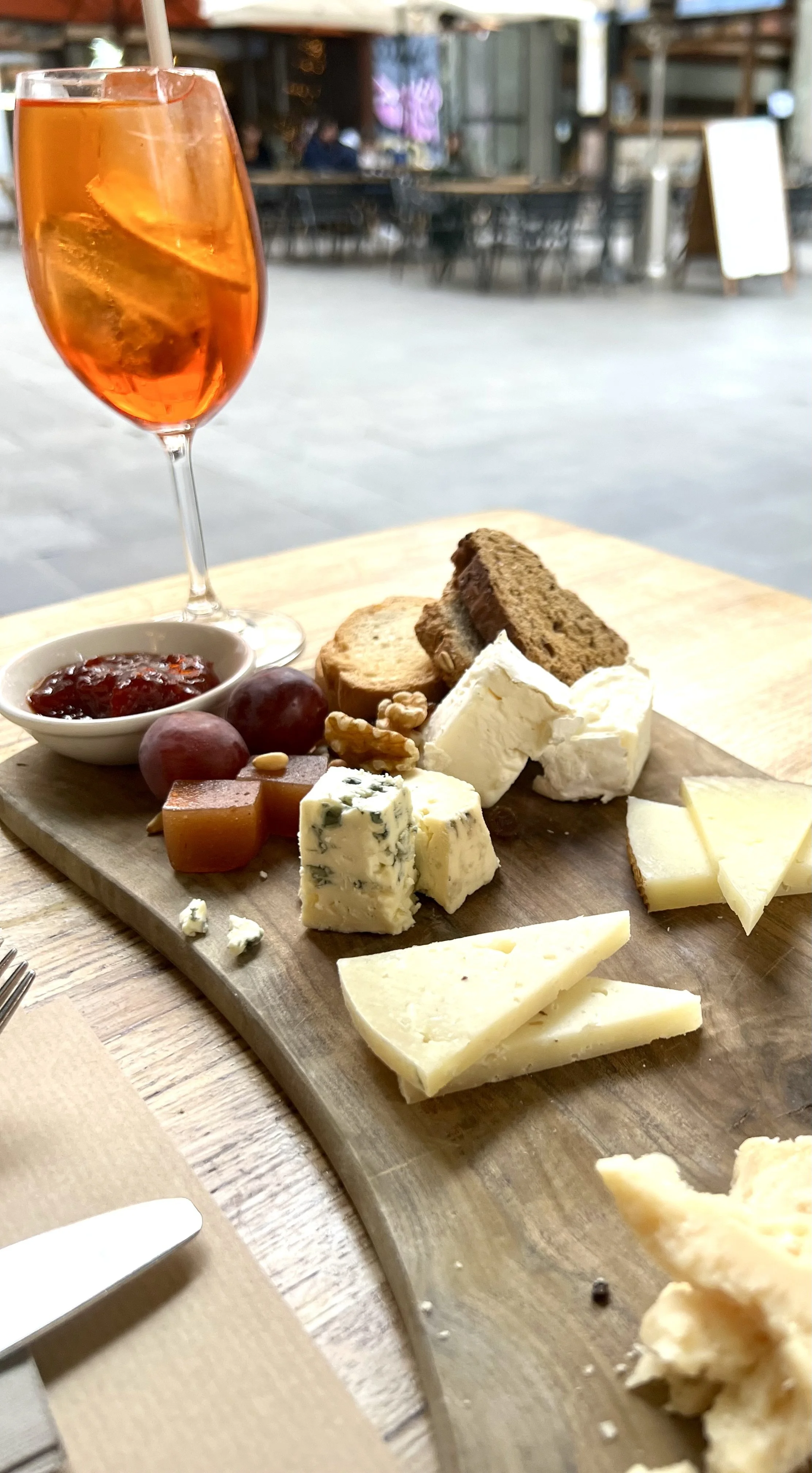 A cheese platter with various cheeses, crackers, grapes, and a small bowl of jam, served with an Aperol Spritz at an outdoor restaurant.