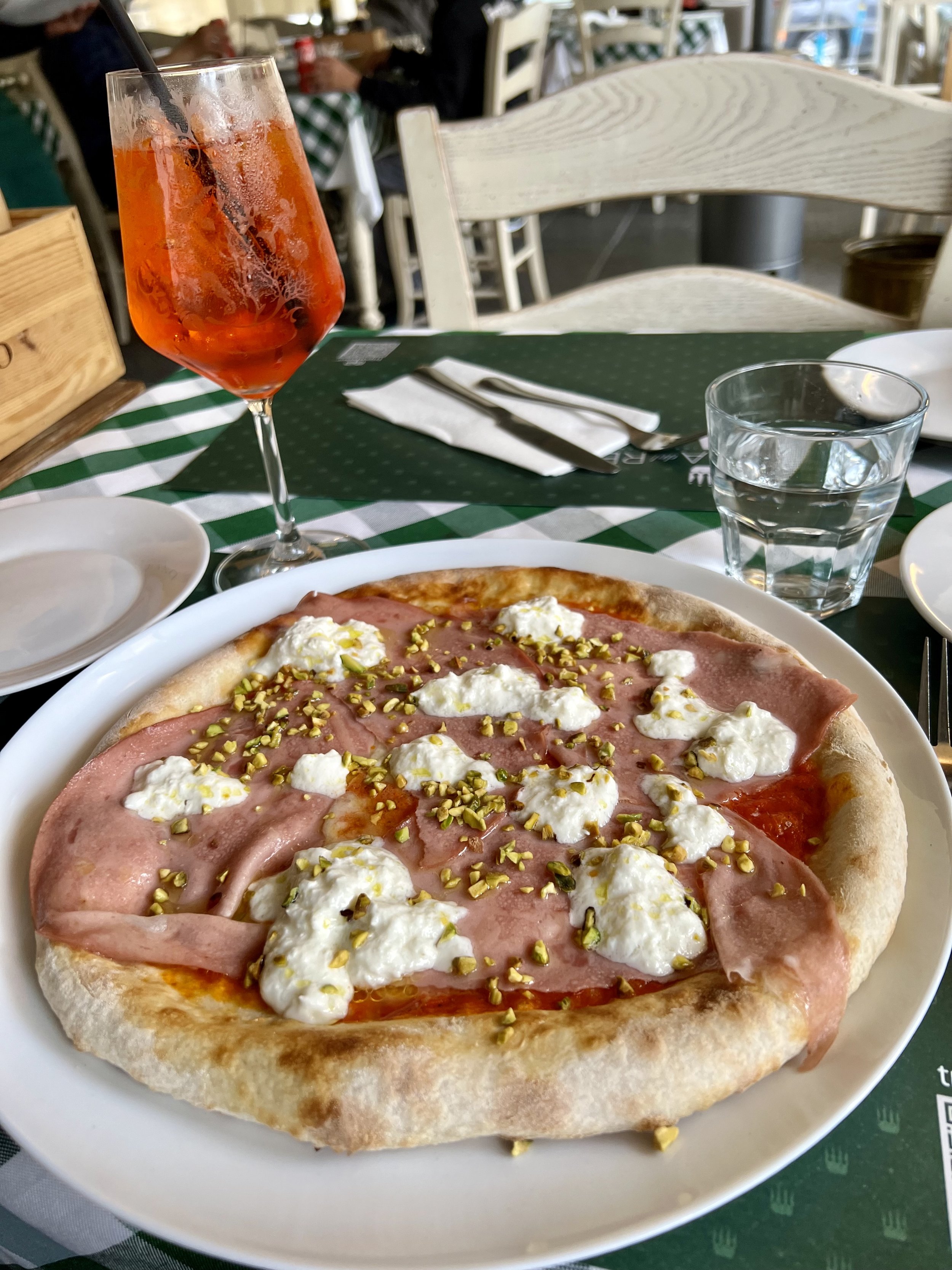 A pizza with mortadella, dollops of burrata cheese, and chopped pistachios on a white plate, with an Aperol Spritz on a table in a restaurant setting.