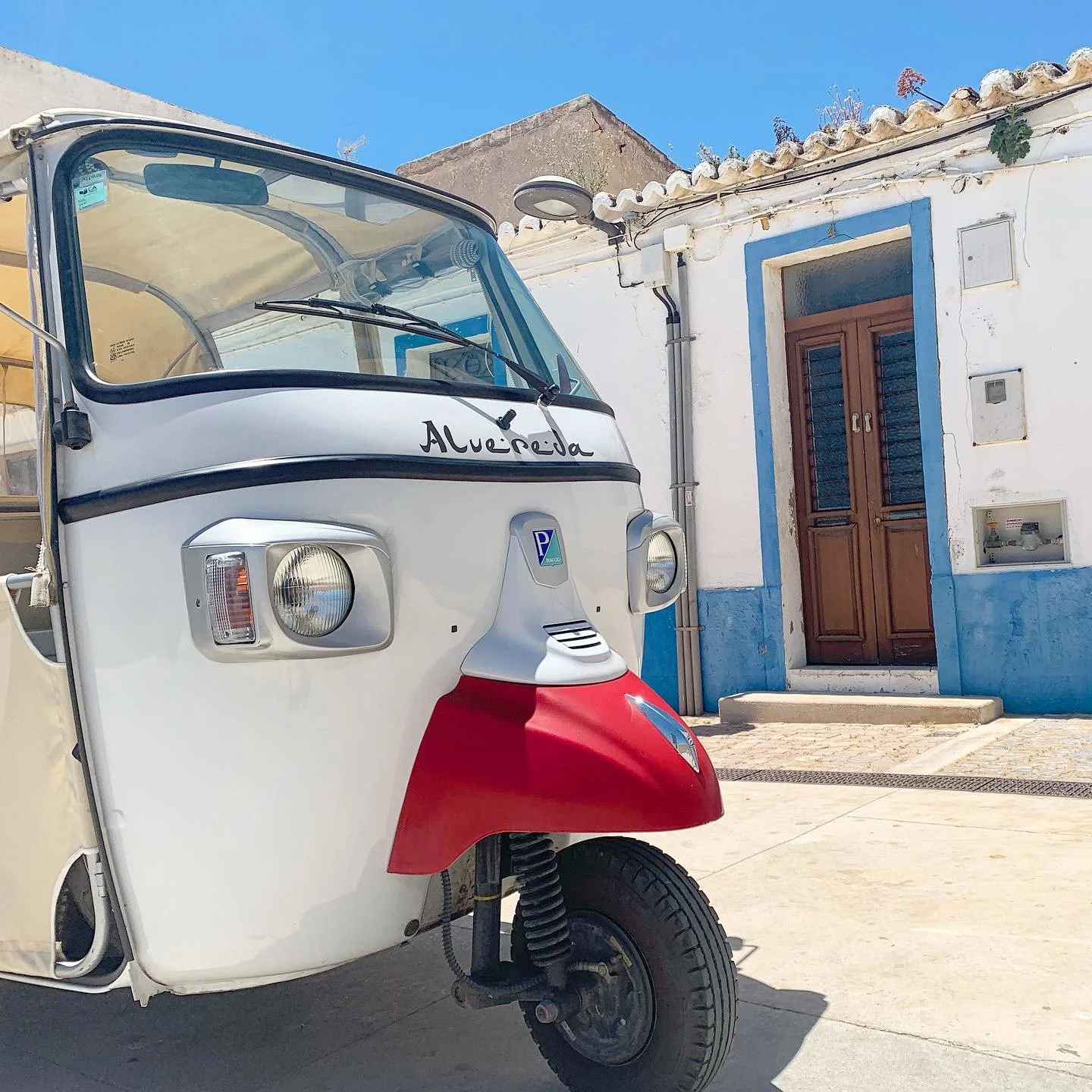 A white, red, and black auto rickshaw parked in front of a white building with a wooden door and blue trim, under a clear blue sky.