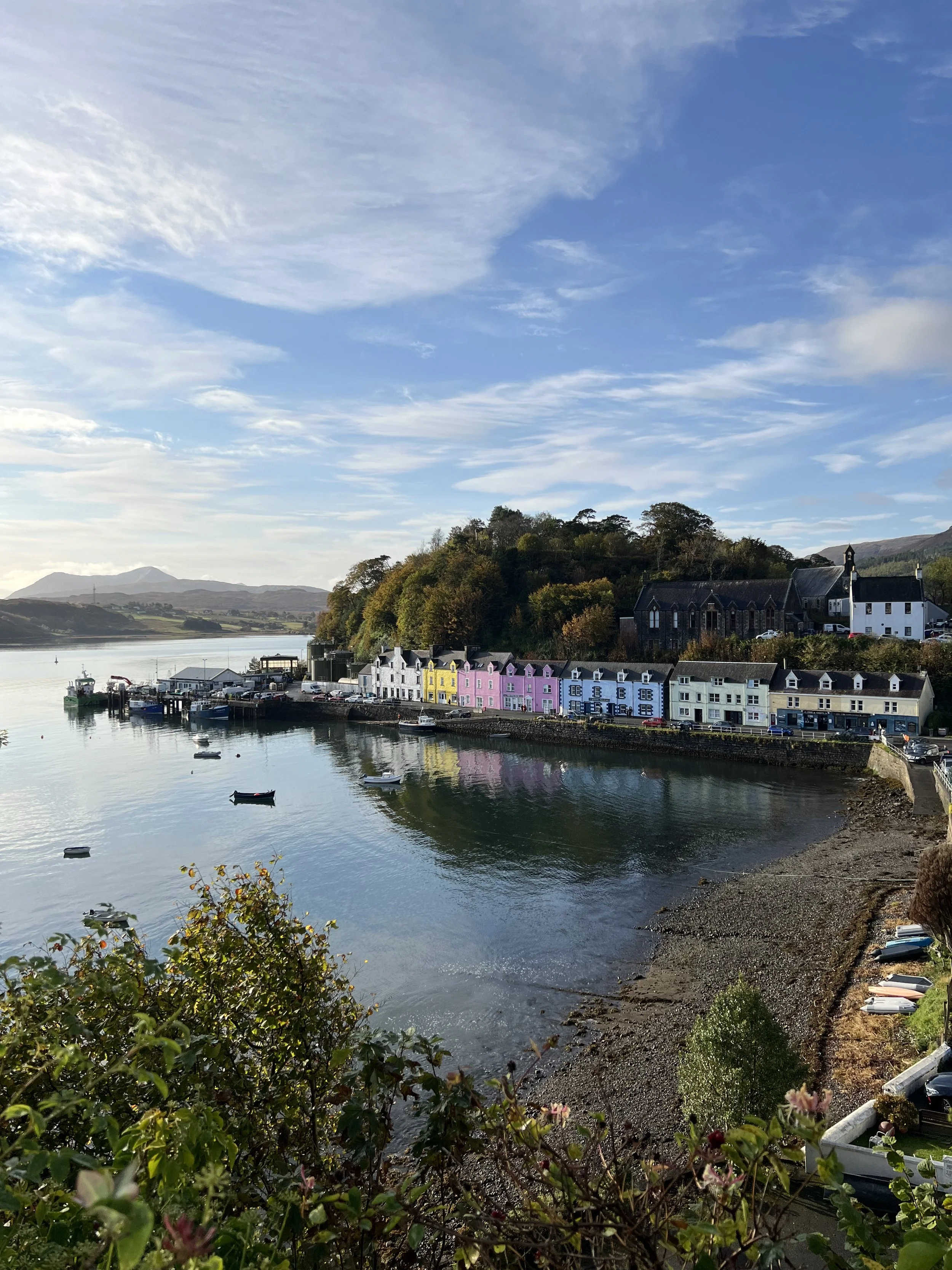 Colorful houses along a waterfront in Portree, Isle of Skye, with boats floating on calm water, a hill with trees in the background, under a partly cloudy sky.