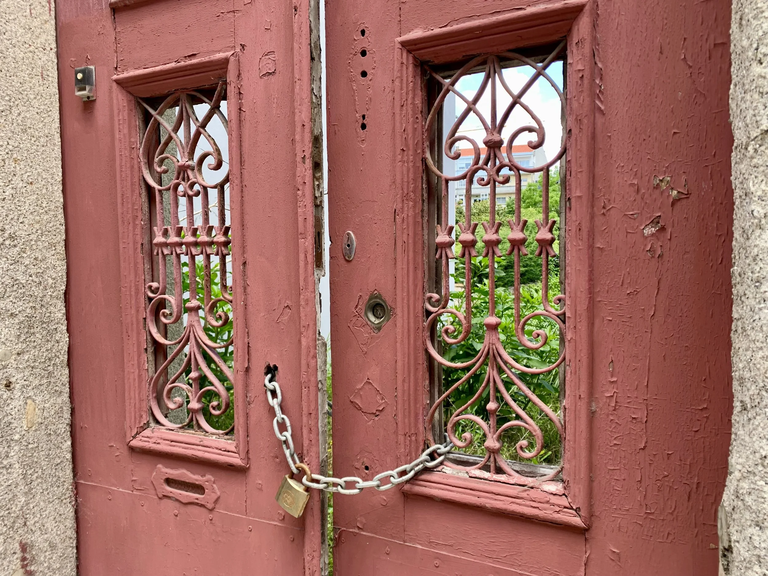 A red wooden gate with ornate metalwork, chained and secured with a padlock, surrounded by concrete walls, with a view of green plants and a building in the background.