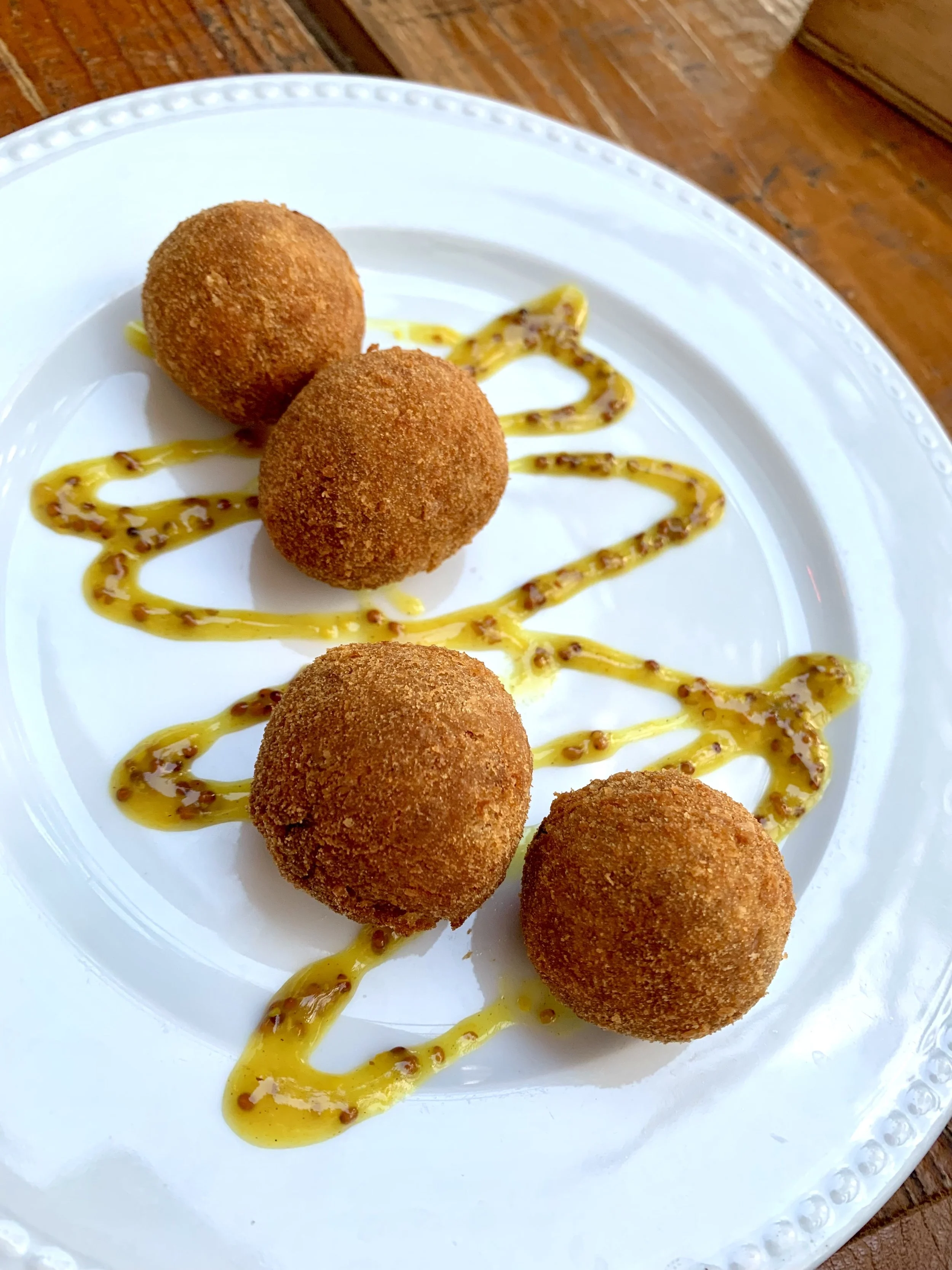 Four golden-brown croquettes on a white plate, garnished with a zigzag drizzle of yellow sauce with mustard seeds, on a wooden table.