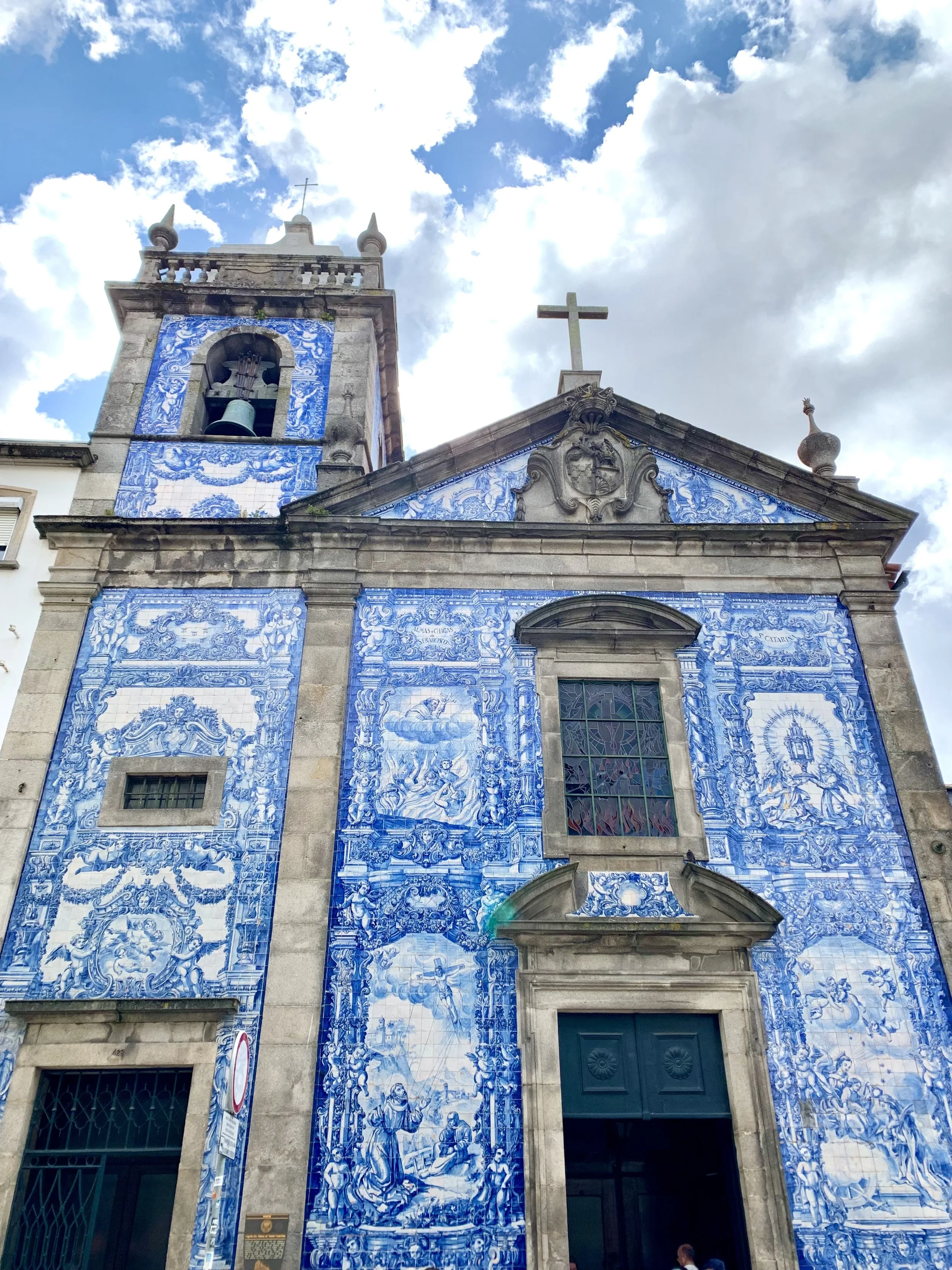 Historical Portuguese church facade decorated with blue and white azulejo tiles, featuring religious scenes, a bell tower, cross, and cloudy sky