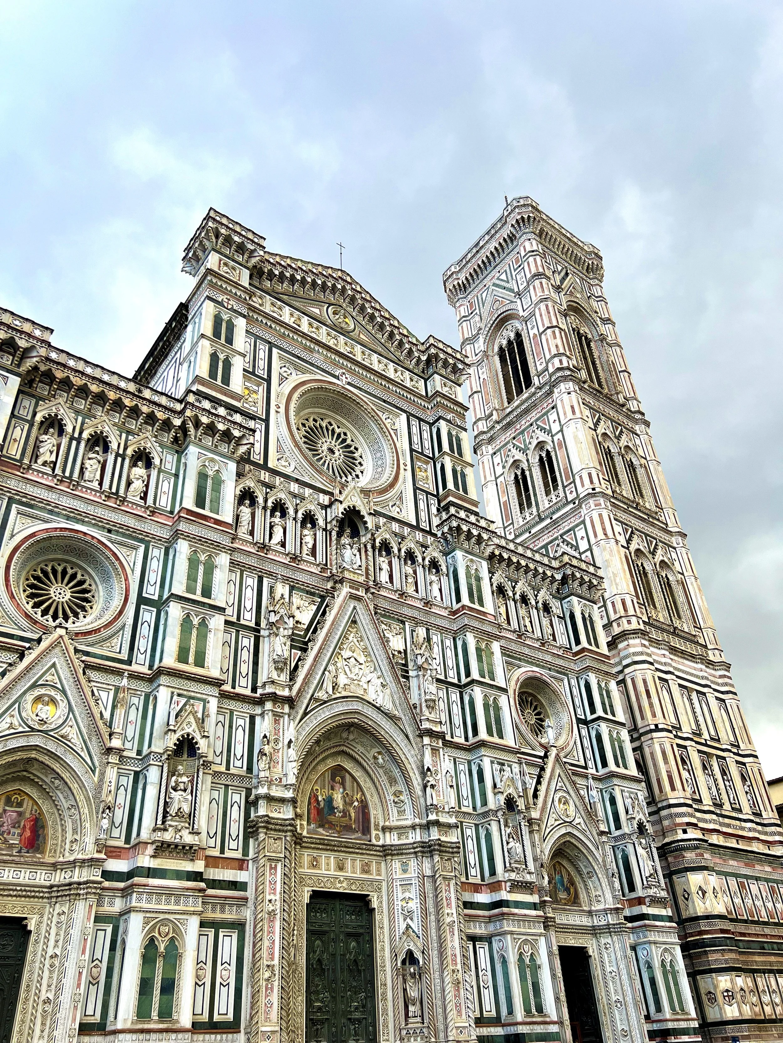 Florence Cathedral, also known as Cattedrale di Santa Maria del Fiore, or The Duomo, showcasing ornate Gothic architecture with detailed marble façade and a tall bell tower, against a cloudy sky.