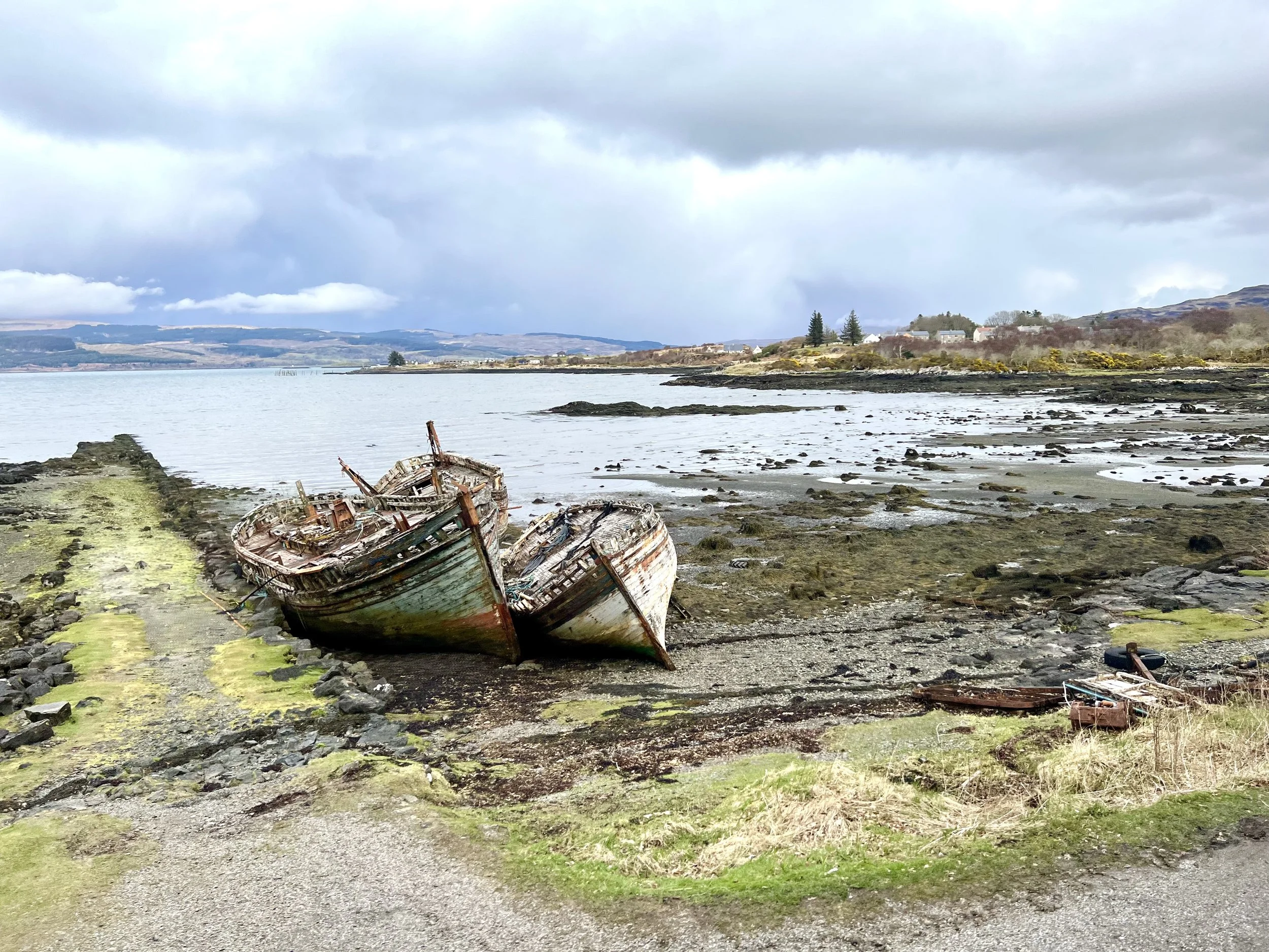 Two decaying, abandoned boats on the rocky shoreline of a body of water, with a distant landscape of hills and cloudy sky in the background.