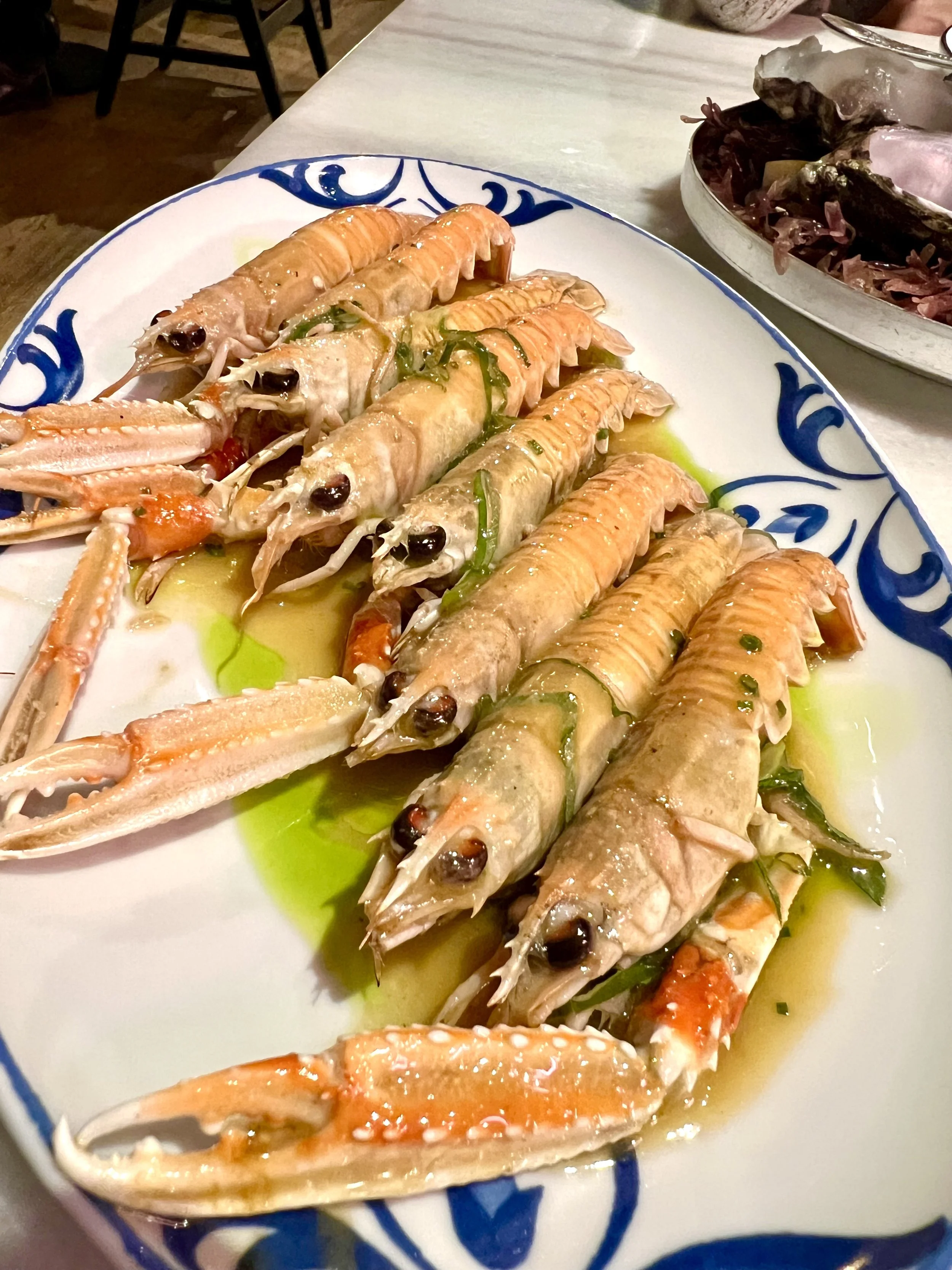 A decorative plate of cooked langoustines garnished with chives, drizzled with olive oil, and served on a white plate with blue patterns.