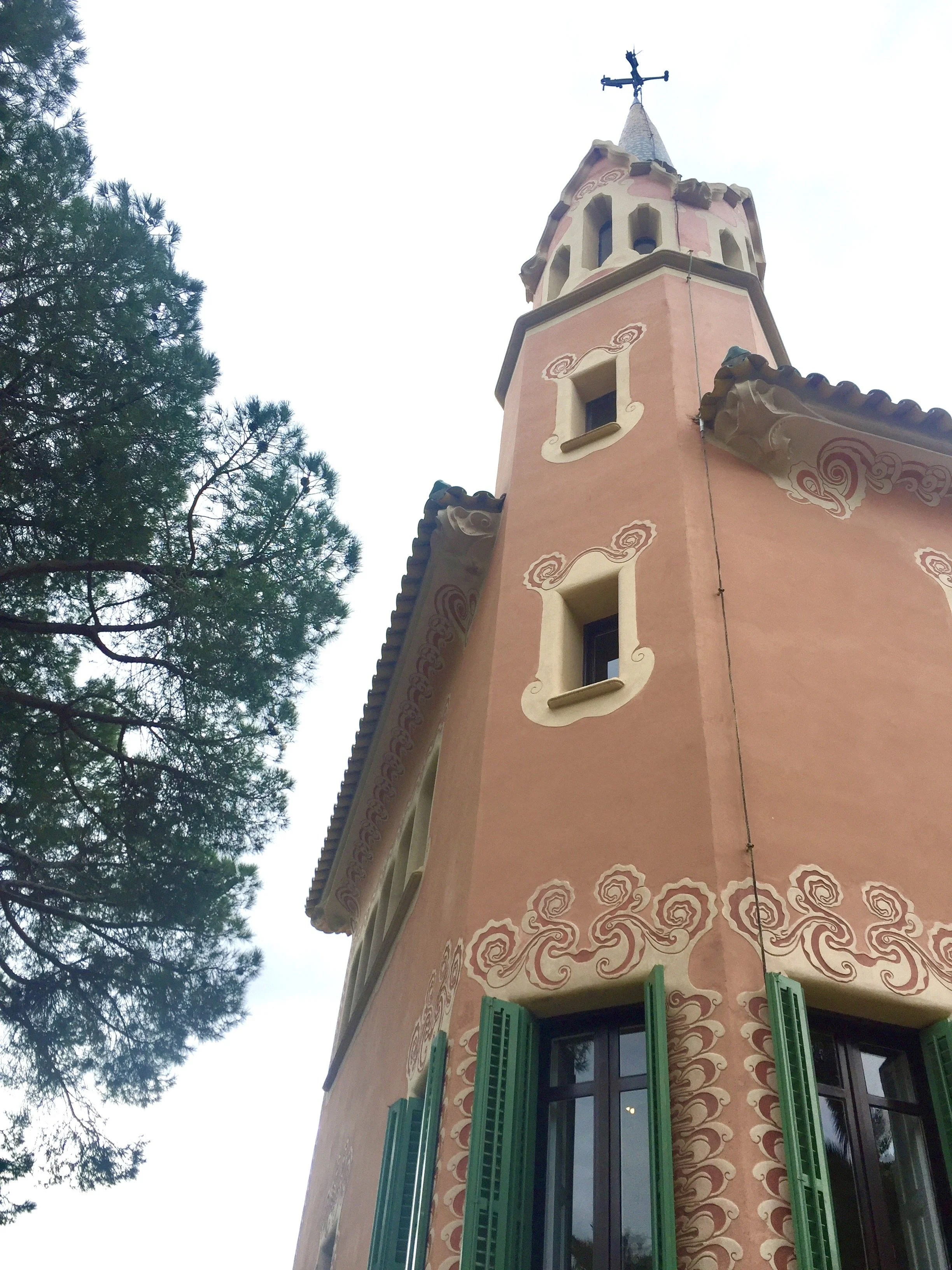 A tall, pink castle-like tower with decorative patterns, green shutters, and a cross at the top, seen from below with a tree on the left.