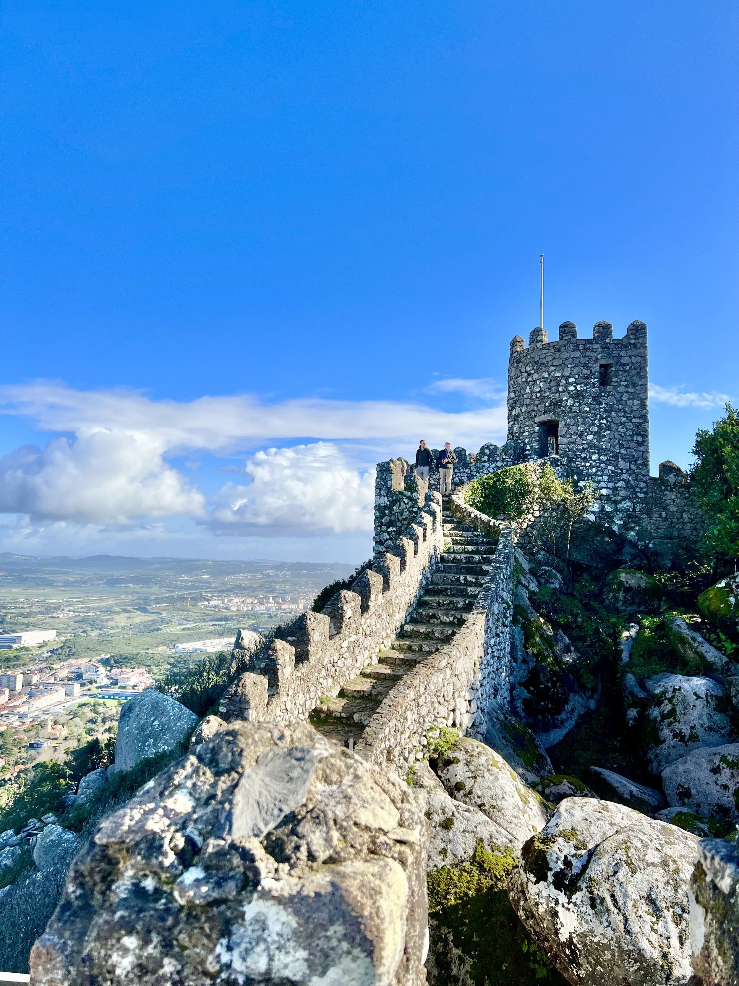 The Moorish Castle in Sintra atop a hill with a staircase, overlooking a city and landscape below, under a blue sky with clouds.