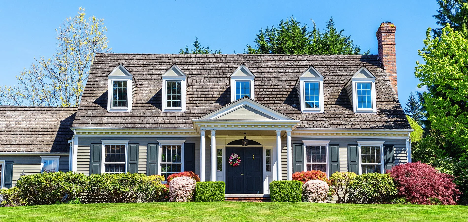 Front view of a traditional house with a brown shingle roof, white siding, black shutters, dormer windows, and a front porch with white columns and a wreath on the door. Well-manicured grass and colorful bushes surround the house, with trees in the background and a clear blue sky.