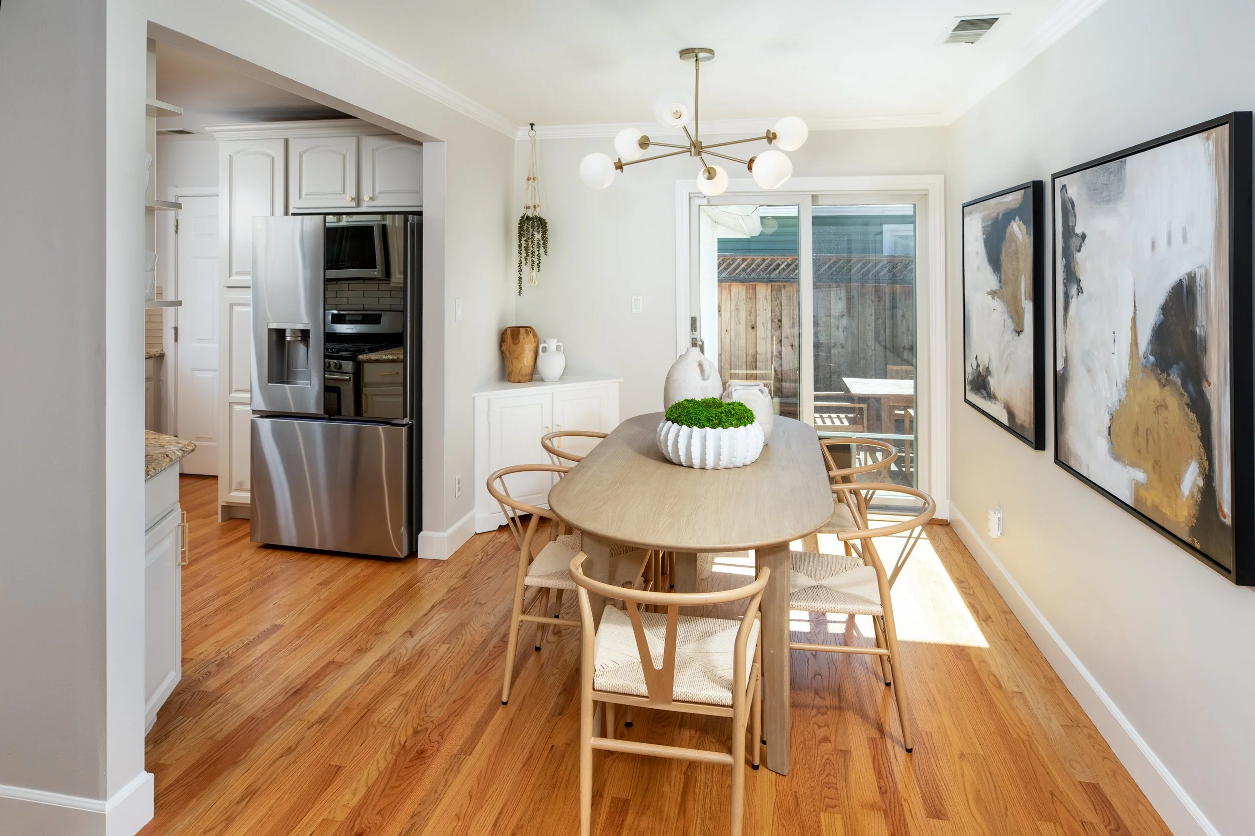 A modern dining area with a wooden table and six matching chairs, a sliding glass door leading outside, a chandelier with multiple globes hanging from the ceiling, and abstract artwork on the wall. The kitchen with a stainless steel refrigerator is p