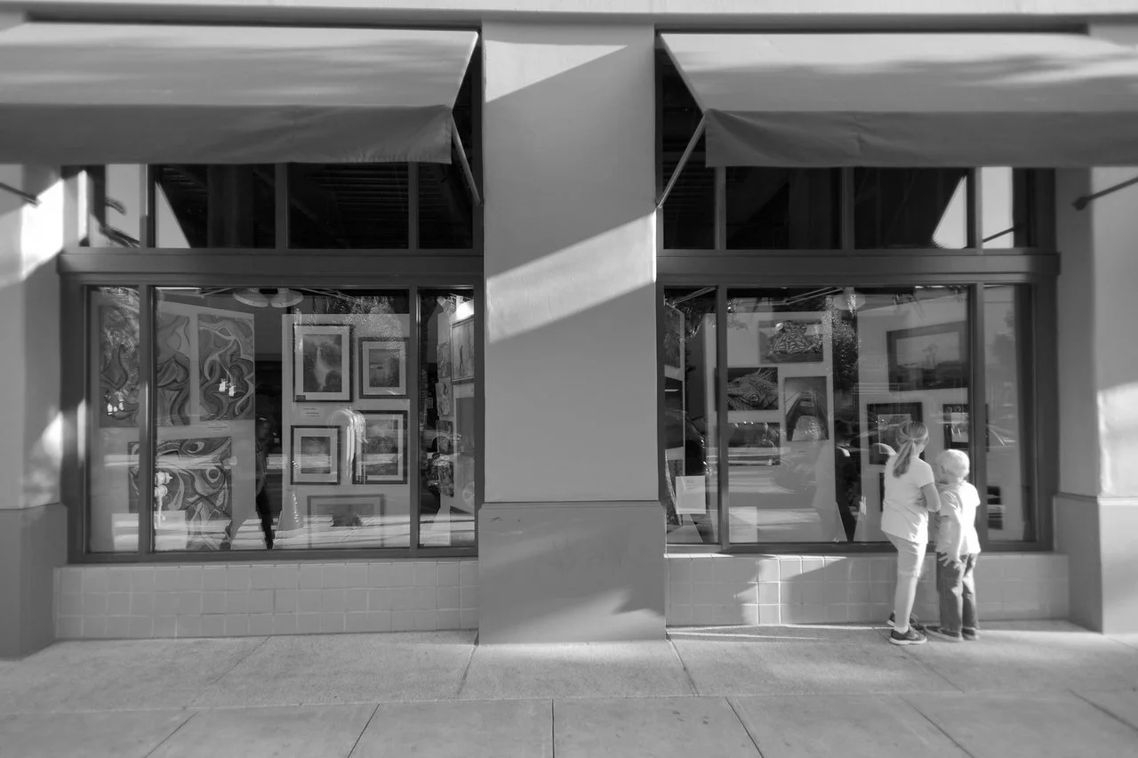 A black and white photo of an art gallery window display, with two children standing together outside, looking at the artwork.