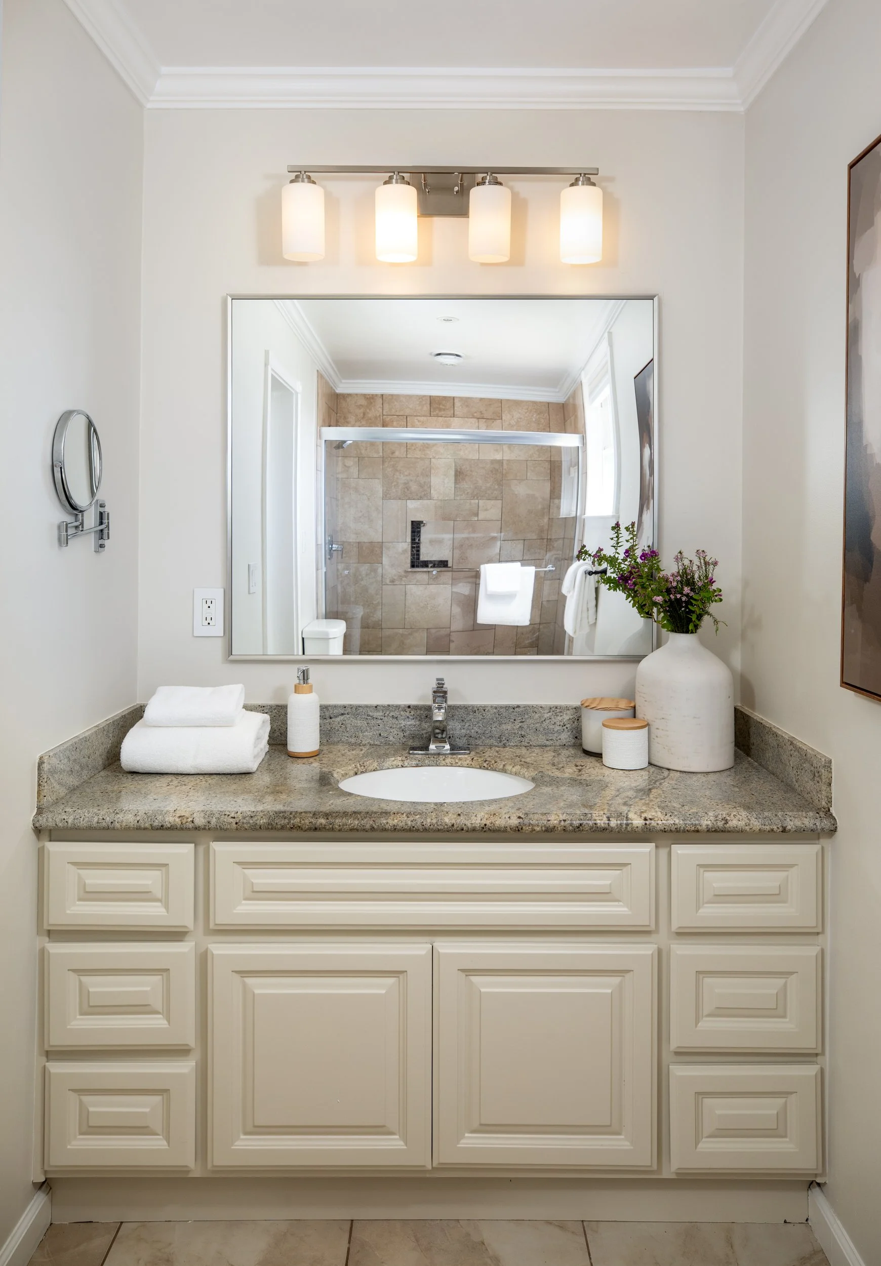Bathroom vanity with a granite countertop, white cabinets, a mirror, and a flower vase, with a view of a shower in the mirror reflection.