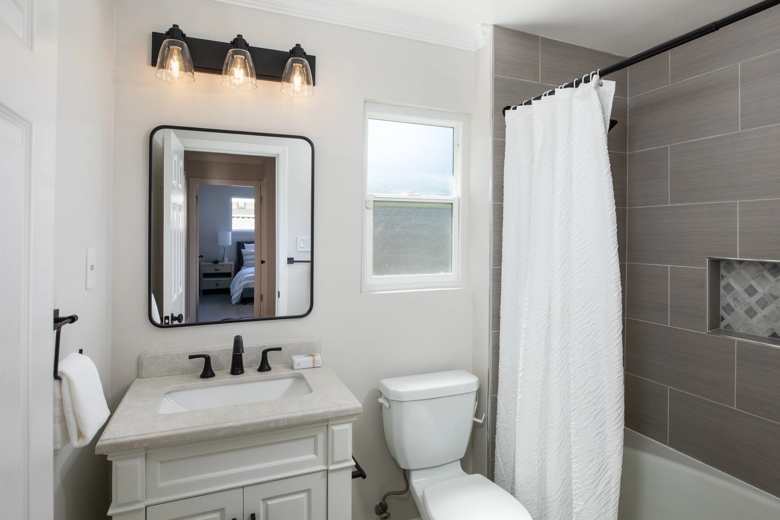 A bathroom with a white vanity sink, a wall-mounted mirror, a toilet, a window, and a shower with a white curtain and gray tiled wall.