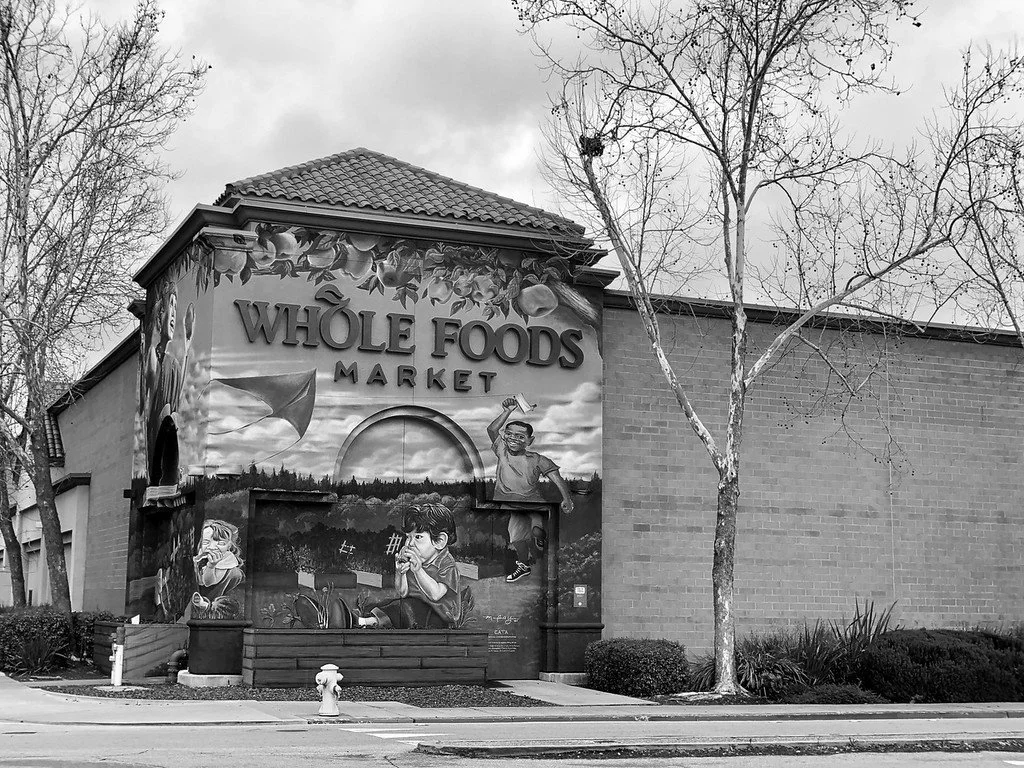 Black and white photo of a Whole Foods Market store with a mural on the front wall, depicting children playing outdoors.