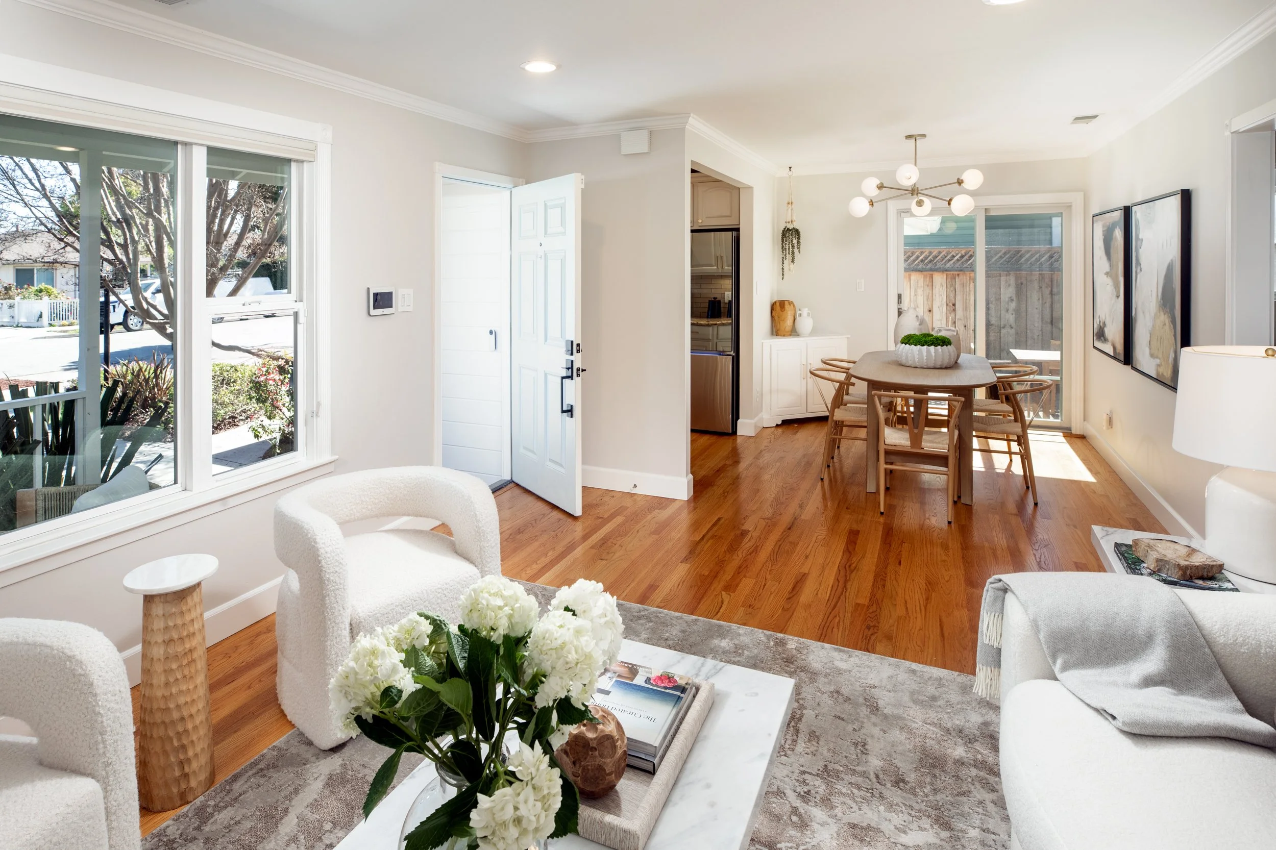 Bright living room with white walls, hardwood floor, large window, white furniture, coffee table with flowers, kitchen and dining area in the background.