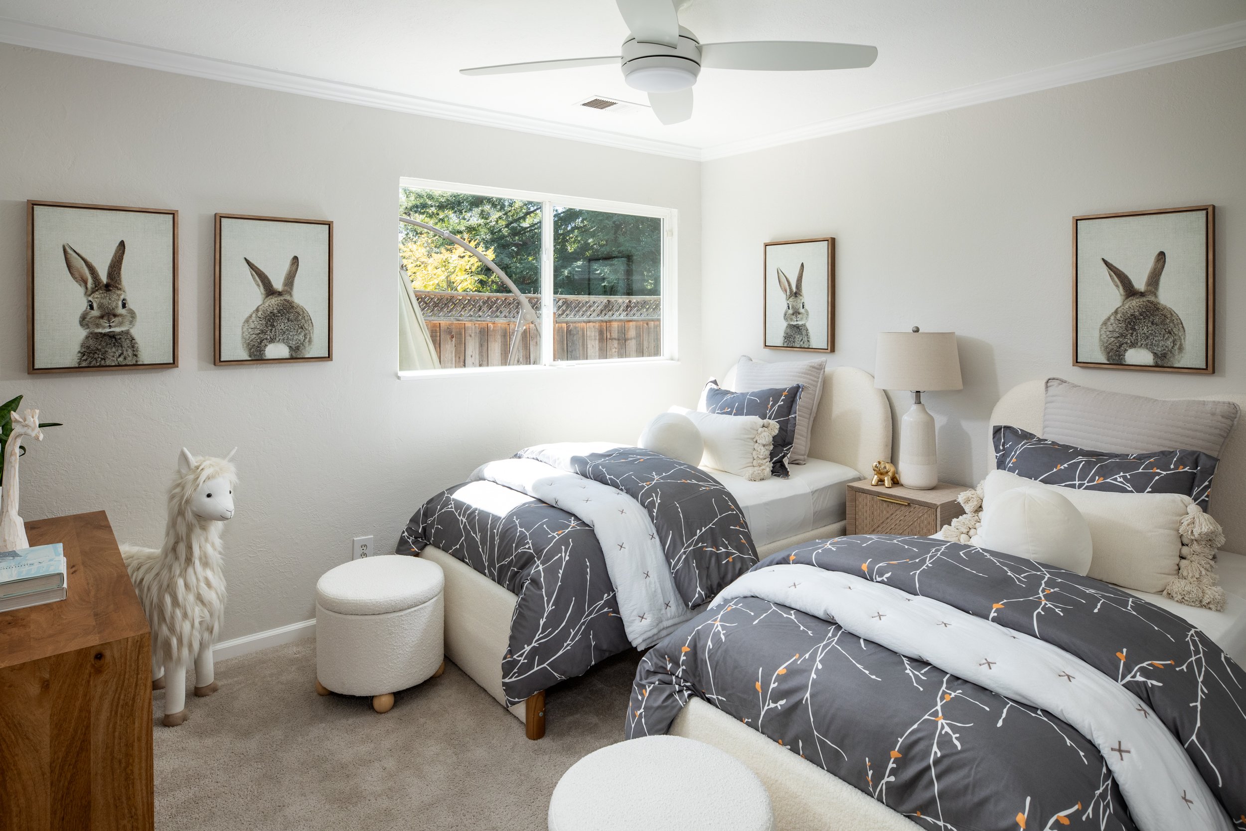 Bedroom with two single beds, black and white bedding with branch pattern, matching pillows and plush toys, lamp, two framed bunny pictures on white wall, window revealing outdoor fence and trees, stuffed unicorn near wooden dresser