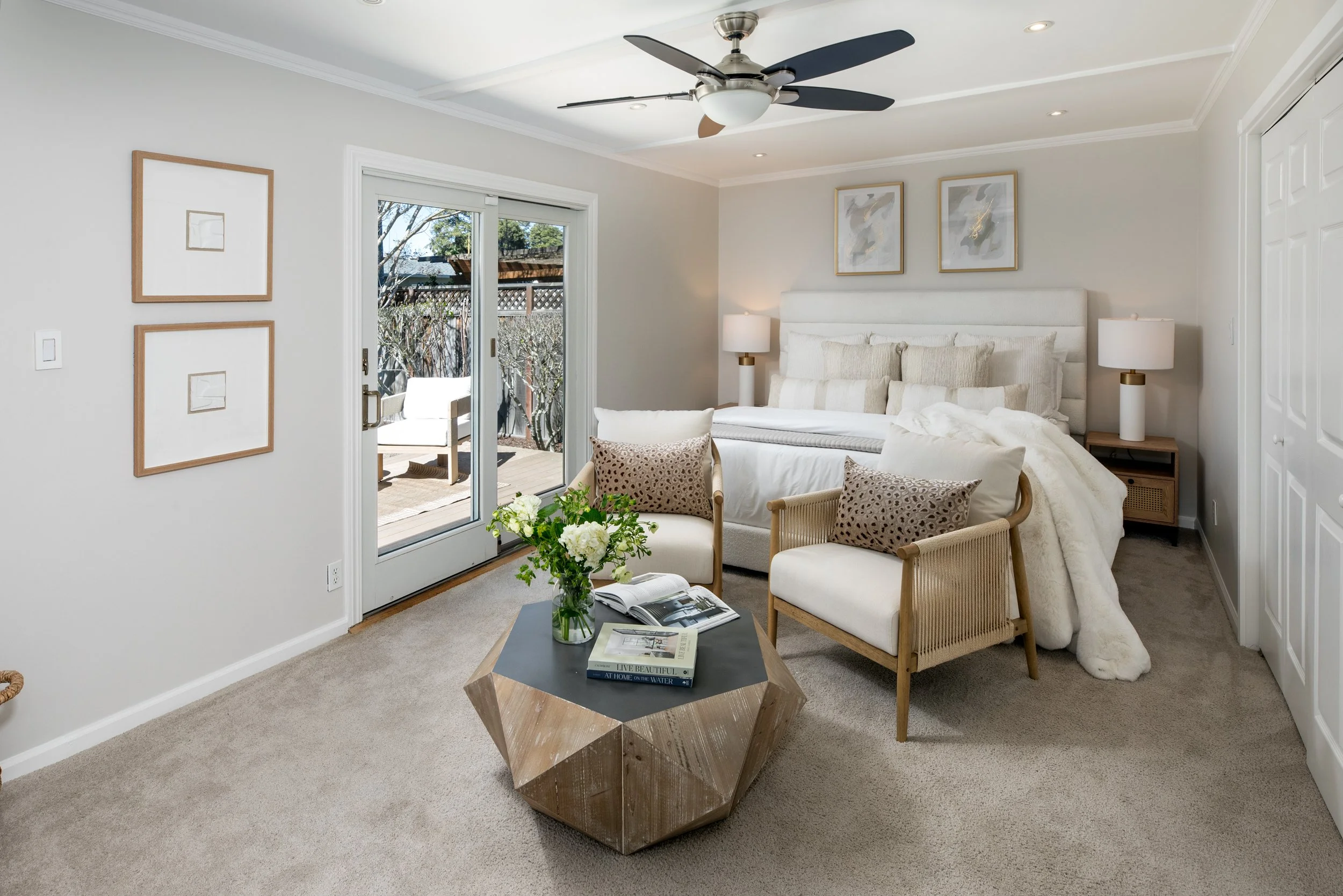 A cozy bedroom with a white upholstered bed, nightstands with lamps, and artwork on the walls. There are two beige armchairs and a geometric coffee table with a vase of flowers. A sliding glass door leads to an outdoor patio with chairs.