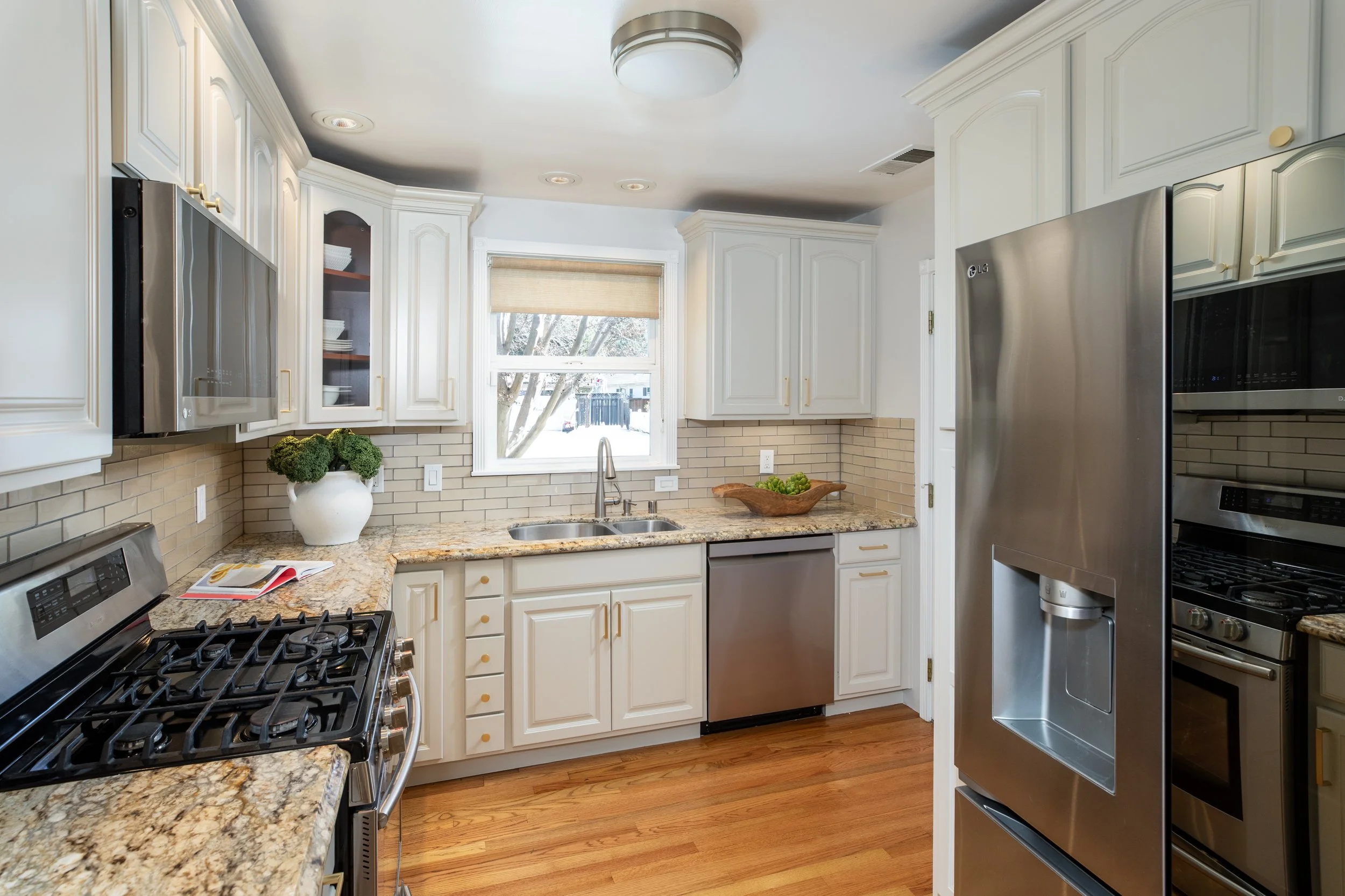 Kitchen with white cabinets, granite countertops, stainless steel appliances, a window with a view outside, and wooden flooring.