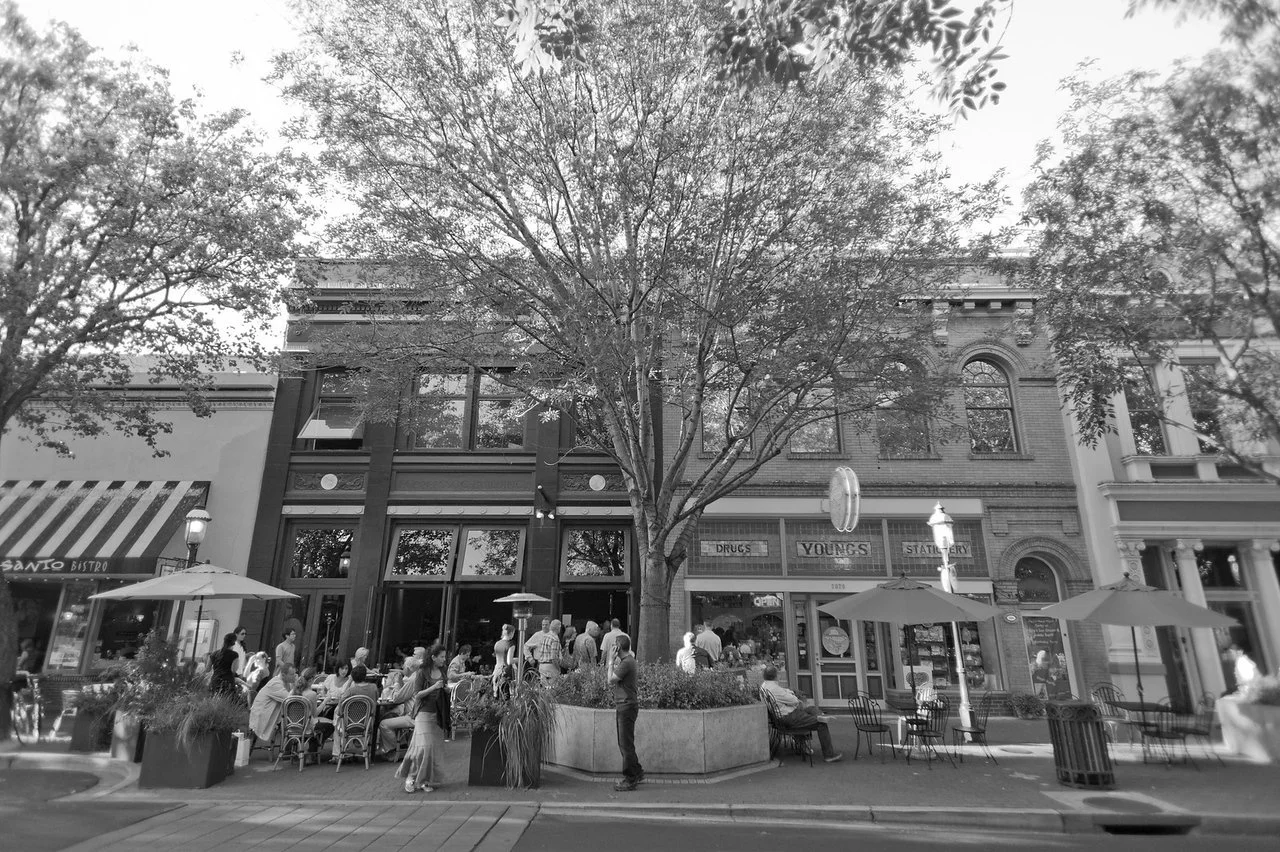 Street scene with a large tree, people sitting at outdoor tables under umbrellas, and a brick building housing a drugstore and stationery shop.