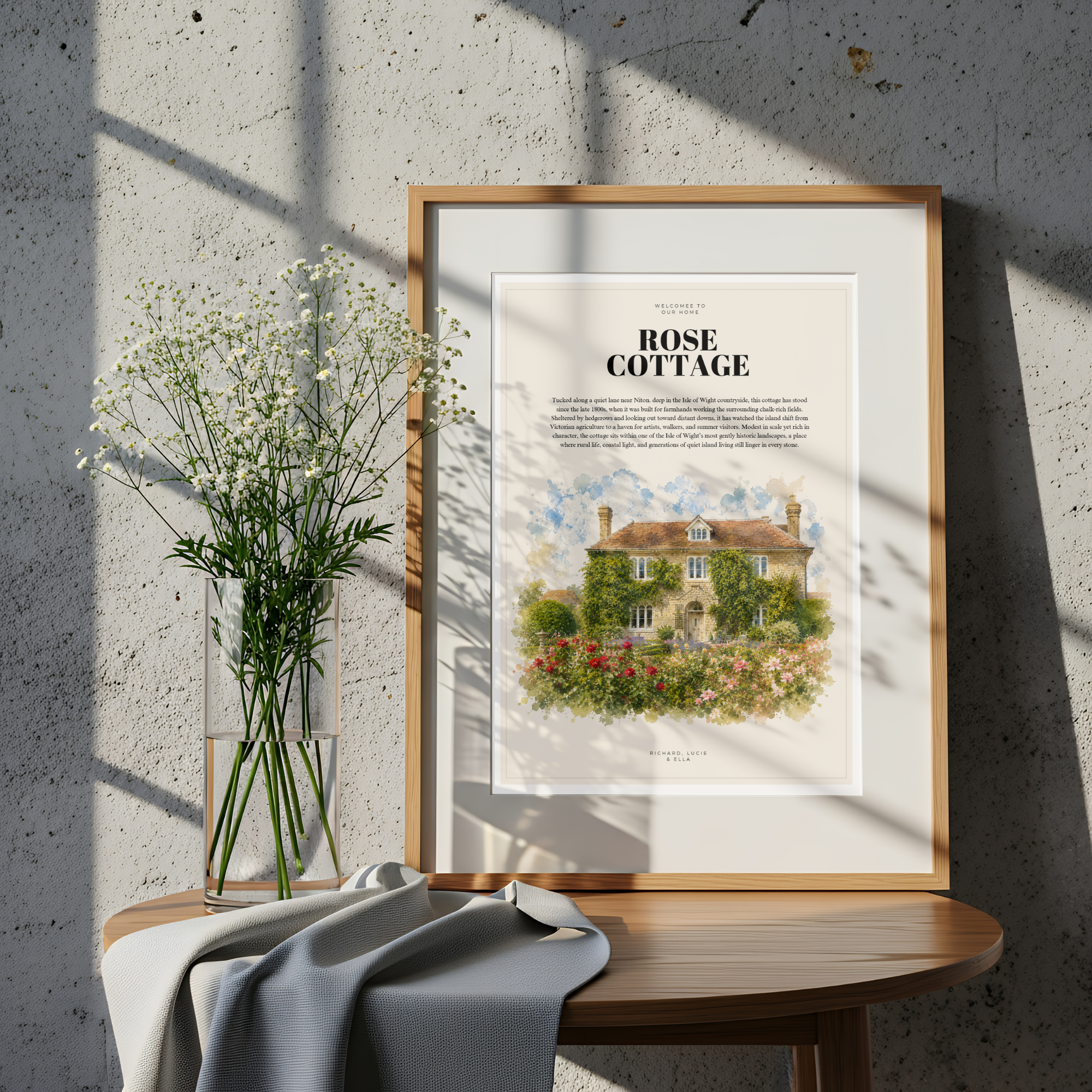 Framed illustration of ‘Rose Cottage’ resting on a round table with a vase of flowers and soft sunlight on a textured wall.