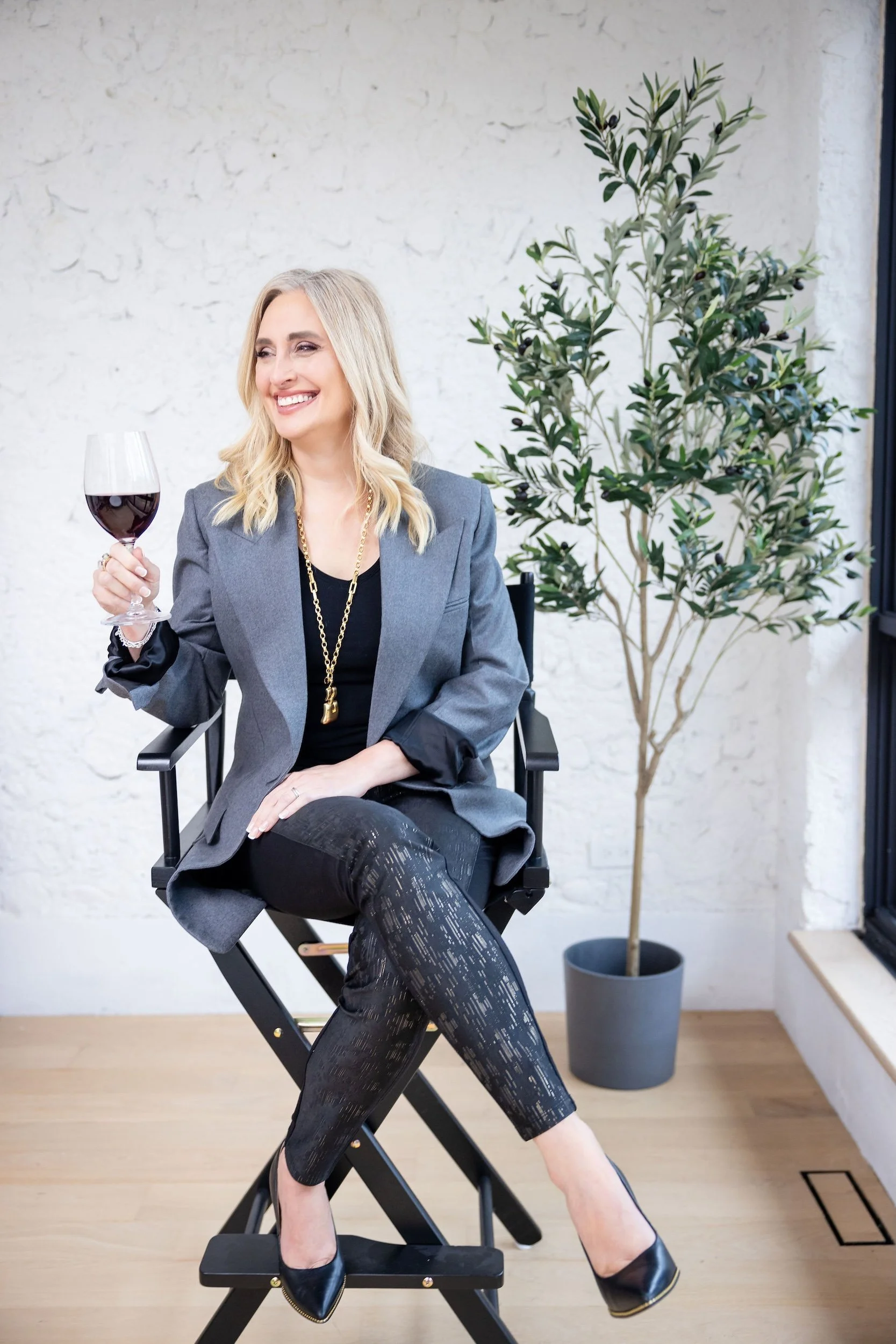 Lauren Brynjelsen sitting on a director's chair, smiling, holding a glass of red wine, wearing a gray blazer, black top, patterned black pants, and black high heels, with a green potted plant and a white wall in the background.